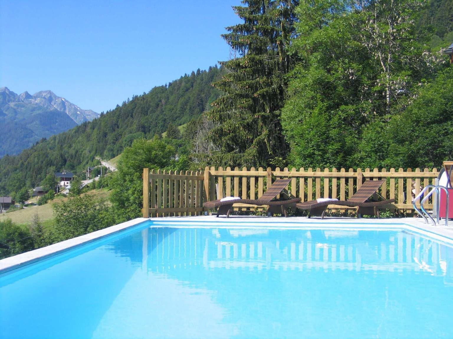 Una piscina con sillones de descanso y un cerco de madera, rodeada de vegetación y montañas al fondo.