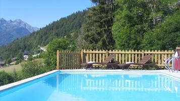 Una piscina con sillones de descanso y un cerco de madera, rodeada de vegetación y montañas al fondo.