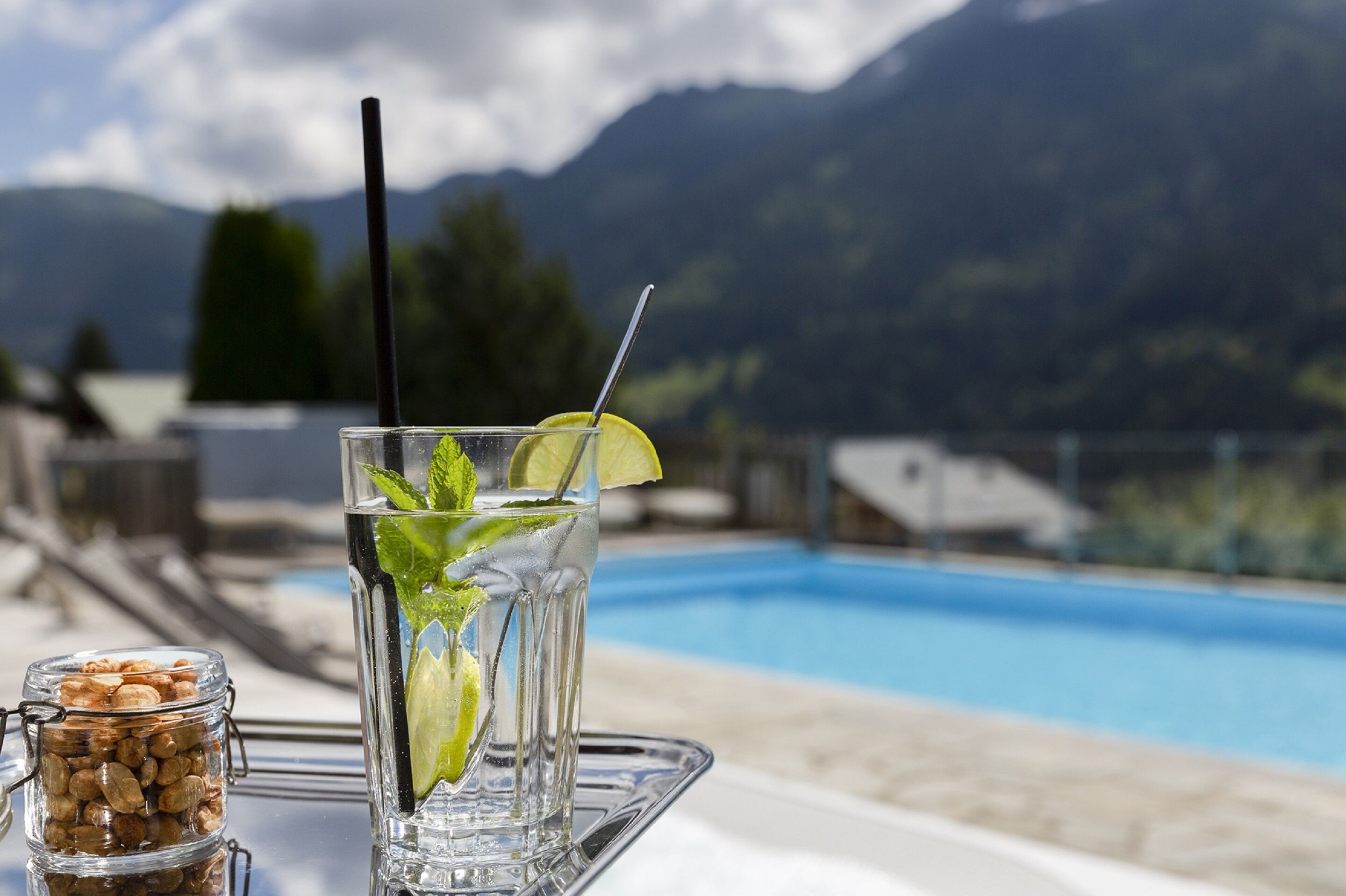 Un verre d’eau à la menthe, garni d’une tranche de citron vert, posé sur un plateau près d’une piscine, avec des montagnes en arrière-plan.