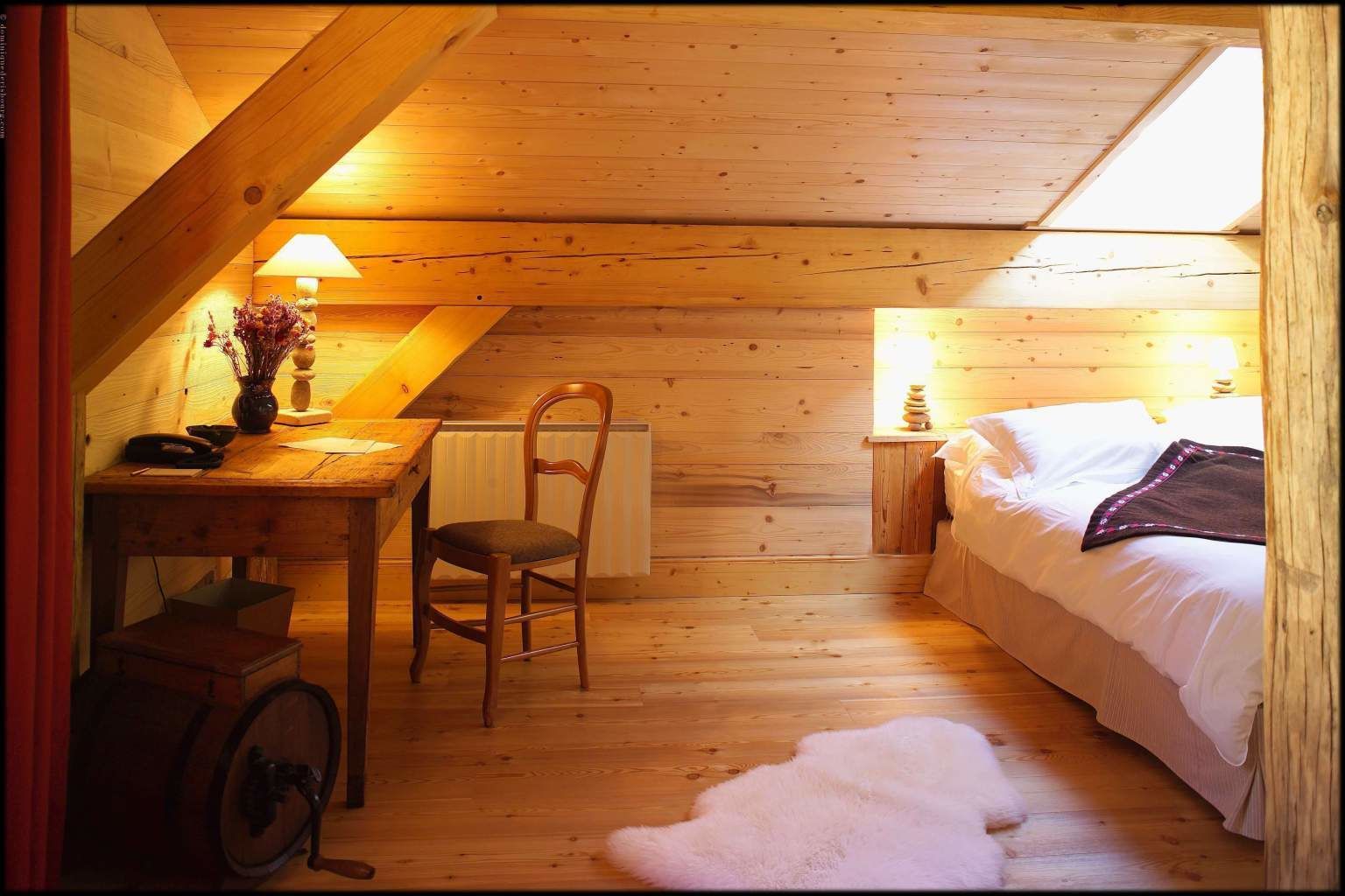 Un dormitorio abrigado en el ático, con suelo de madera, un escritorio con lámpara, una silla, una cama con almohadas y un radiador.