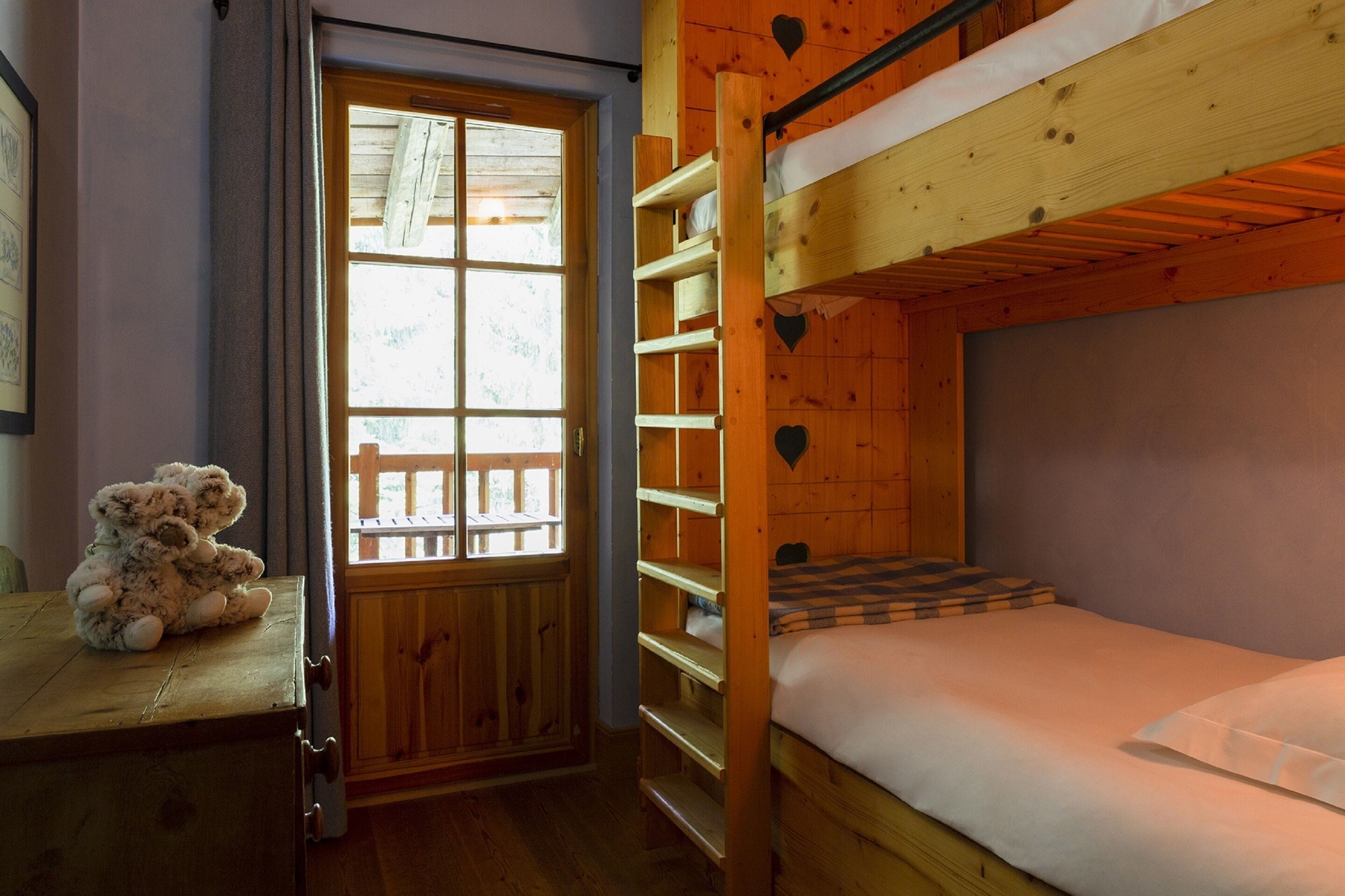 A bunk bed room with a wooden door and window, a teddy bear on a table, and a checkered bedspread.