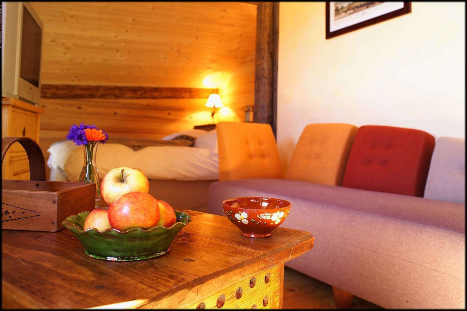 A cozy room with a wooden bed, a small table with a bowl of fruit, and a lamp.