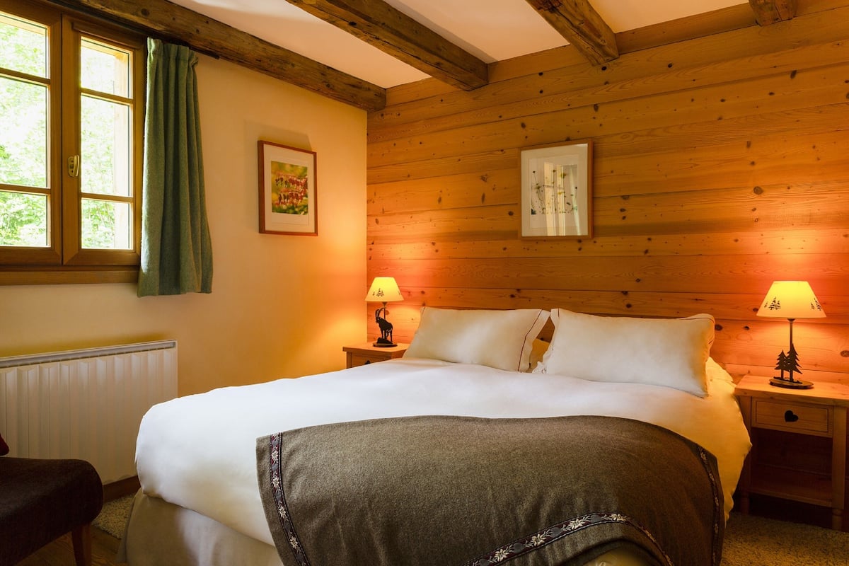 A cozy bedroom with a wooden ceiling, a large bed with white linens, two bedside lamps, and a window with a green curtain.