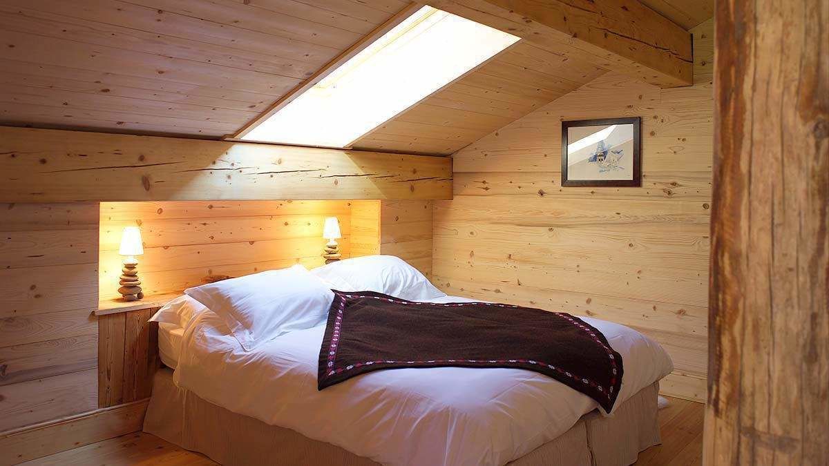 Un dormitorio de cabaña de madera acogedor, con tragaluces, una cama con sábanas blancas, una lámpara de noche y un cuadro en la pared.