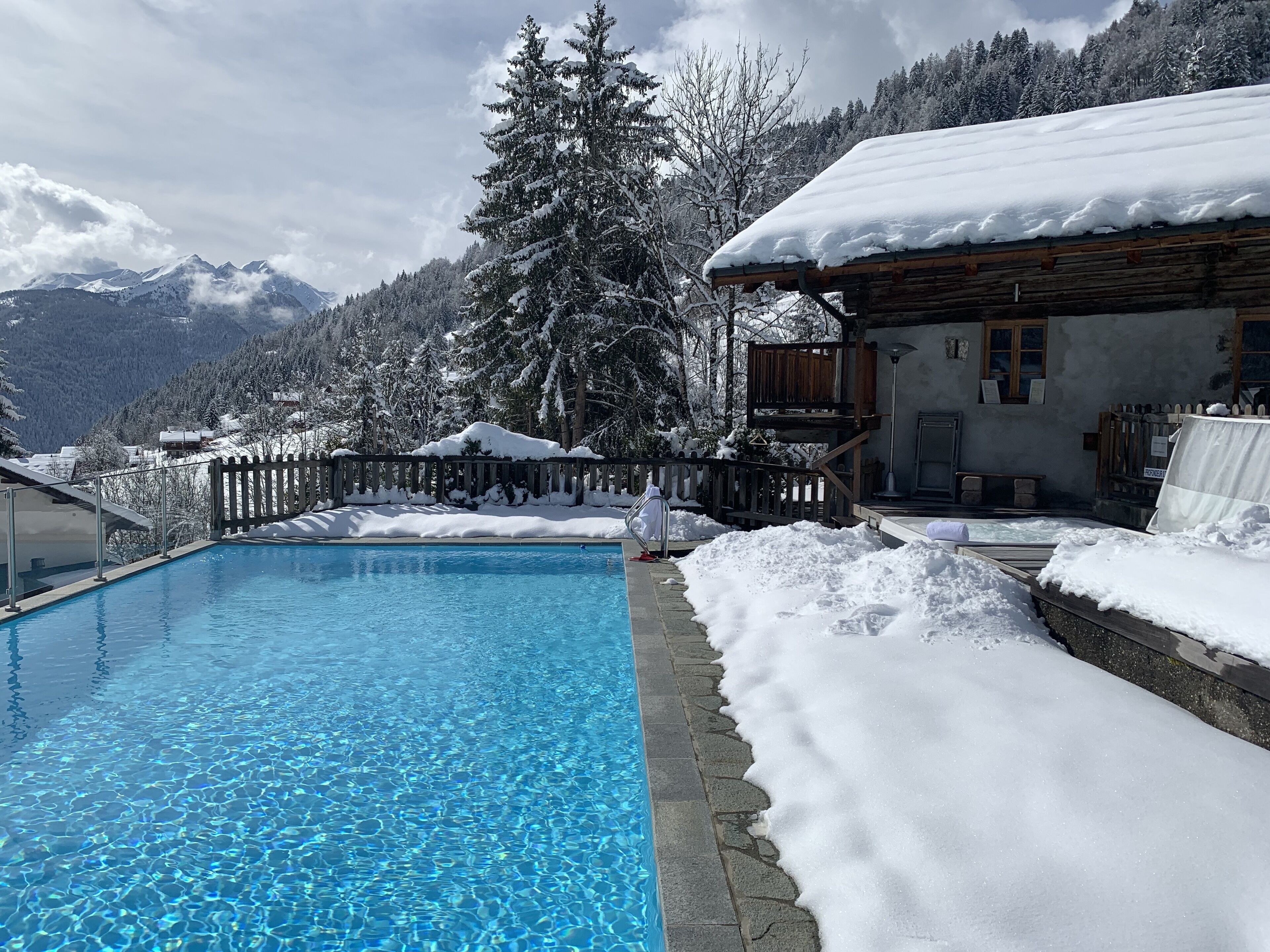 A swimming pool with clear blue water, surrounded by snow-covered ground and a wooden building with a balcony.