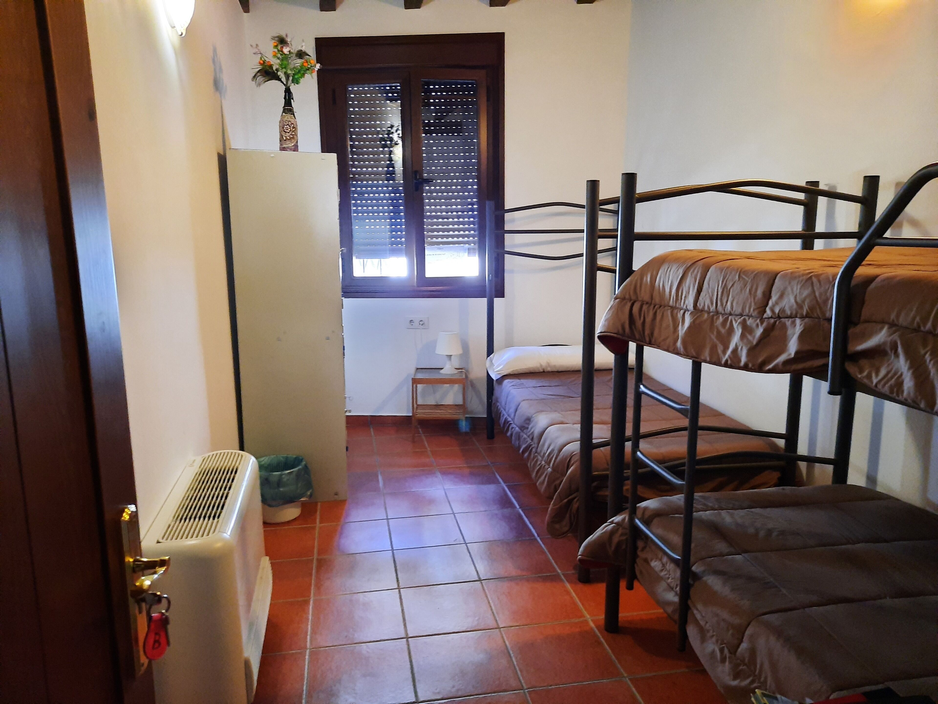 Une chambre de dortoir avec des lits superposés, un radiateur, une fenêtre avec des stores et une petite table de chevet avec une lampe.