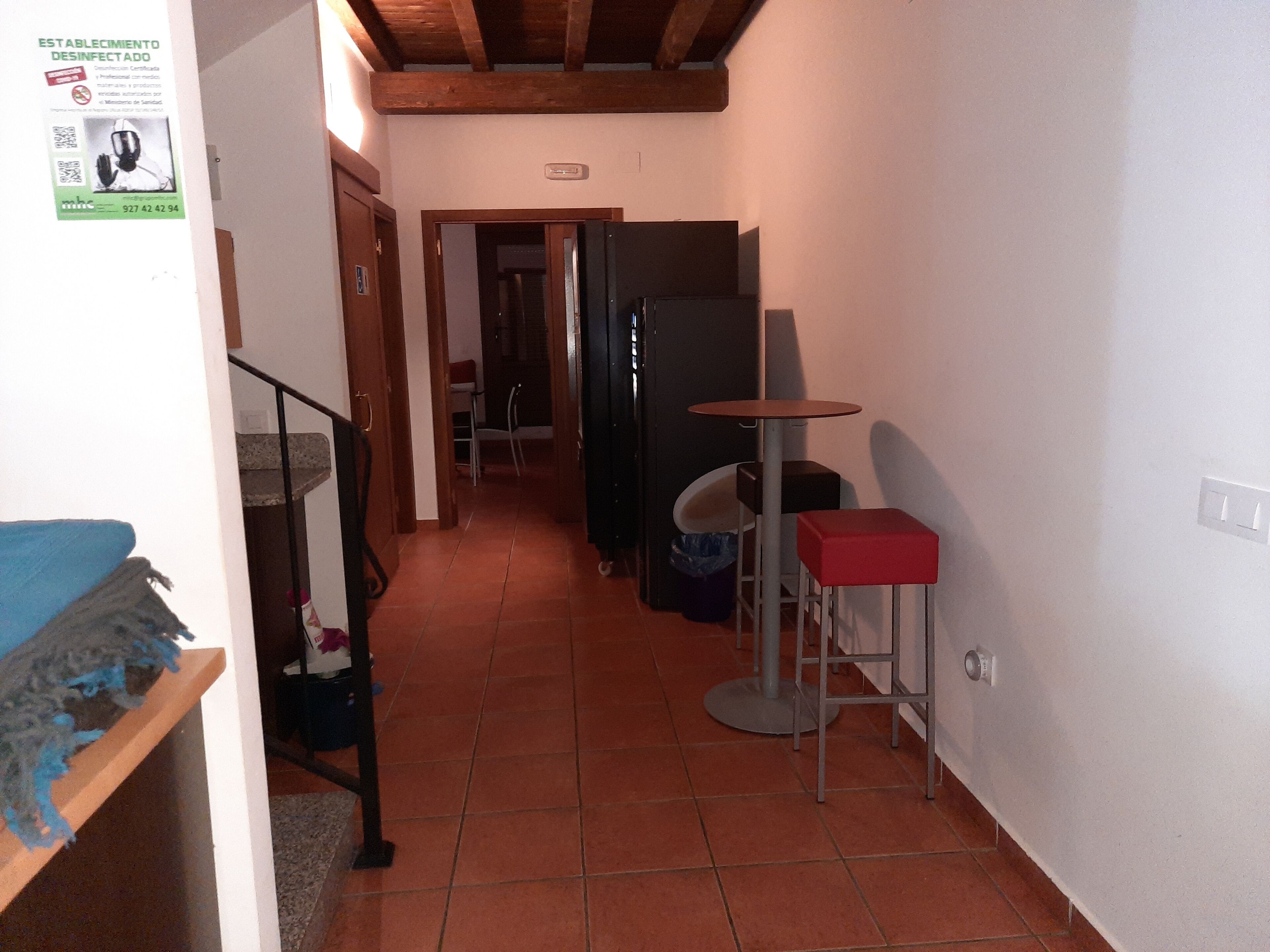 A narrow hallway with a tiled floor, a small table with a red stool, a black refrigerator, and a door leading to another room.