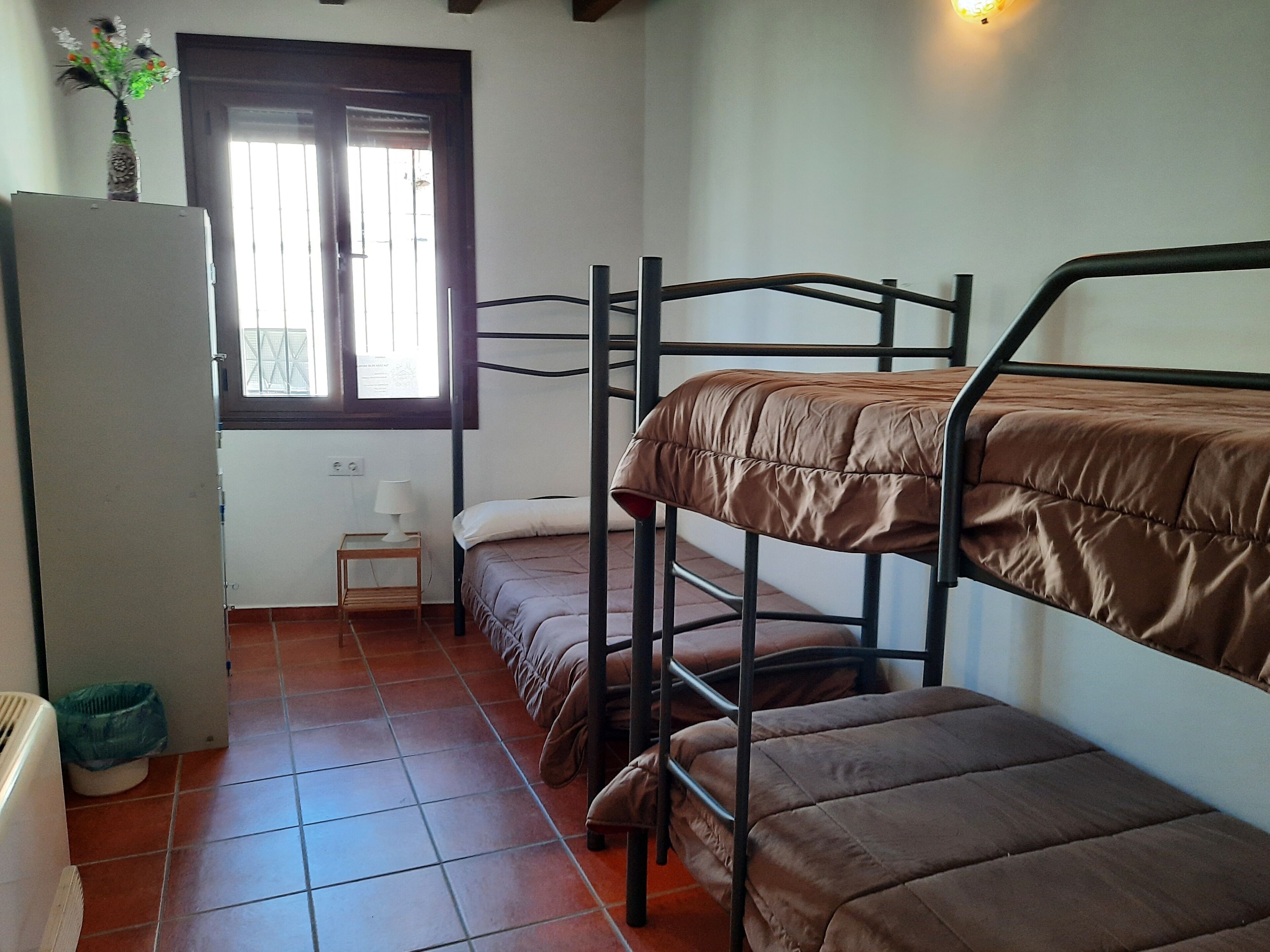 A dormitory room with bunk beds, a small table with a lamp, a window with blinds, and a refrigerator.