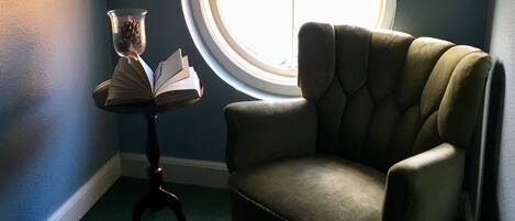Quarto de canto com janela redonda, uma cadeira e uma pequena mesa com um livro e um vaso.