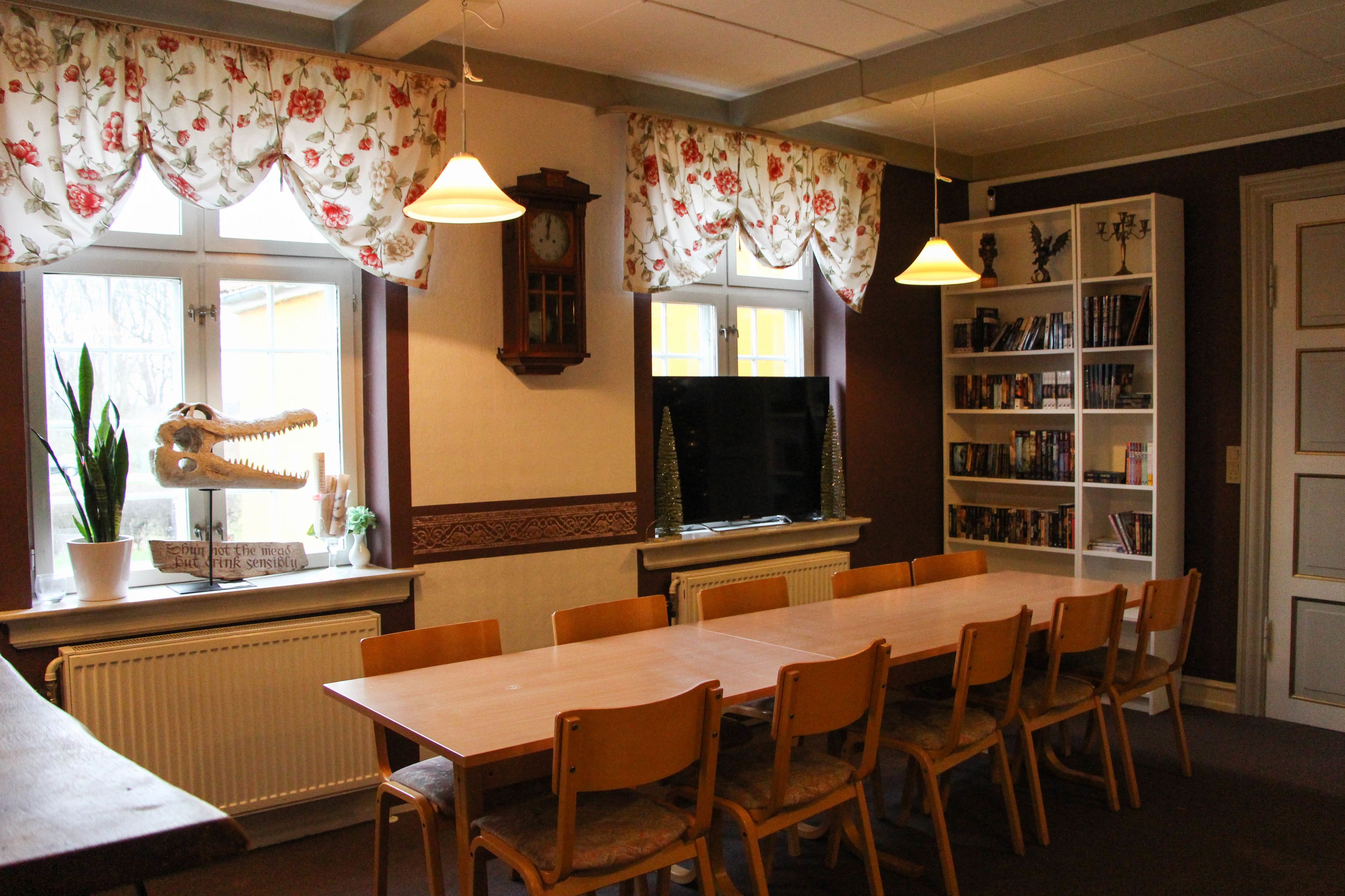 A dining room with a long table, chairs, a clock, a bookshelf, and a window with curtains.