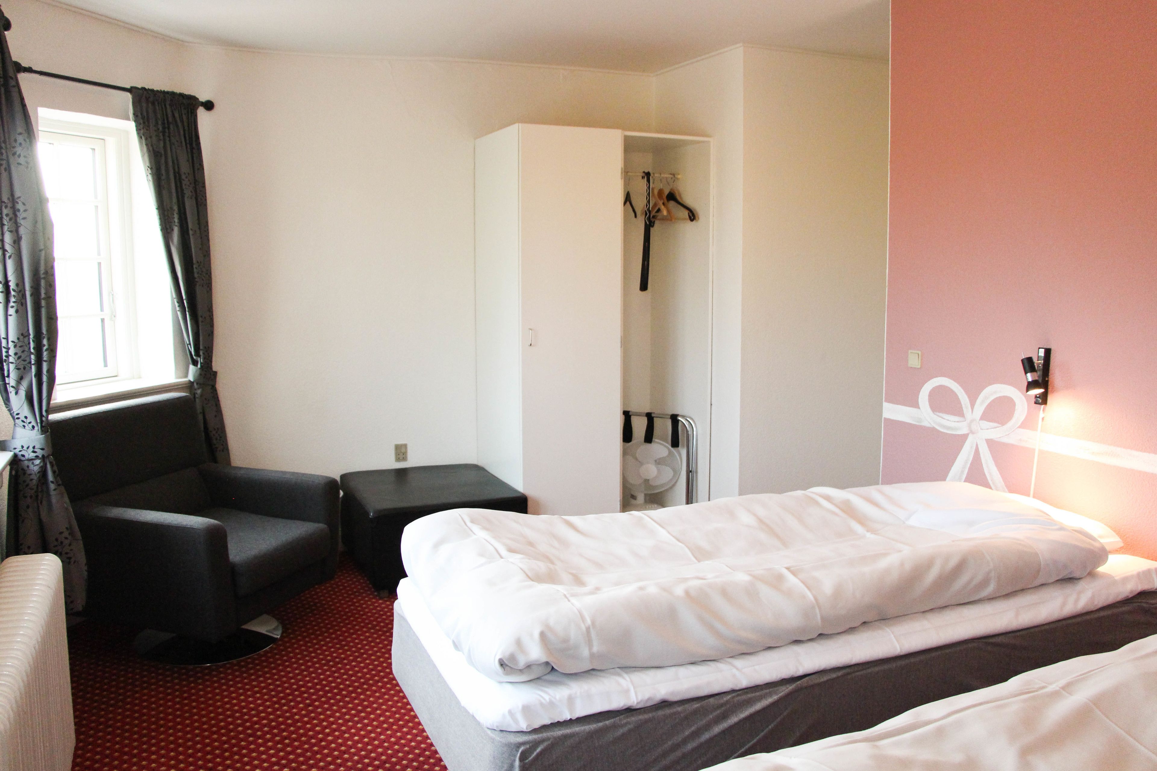 Una habitación de hotel con cama, una silla, una mesita, un armario y un ventanal con cortinas.