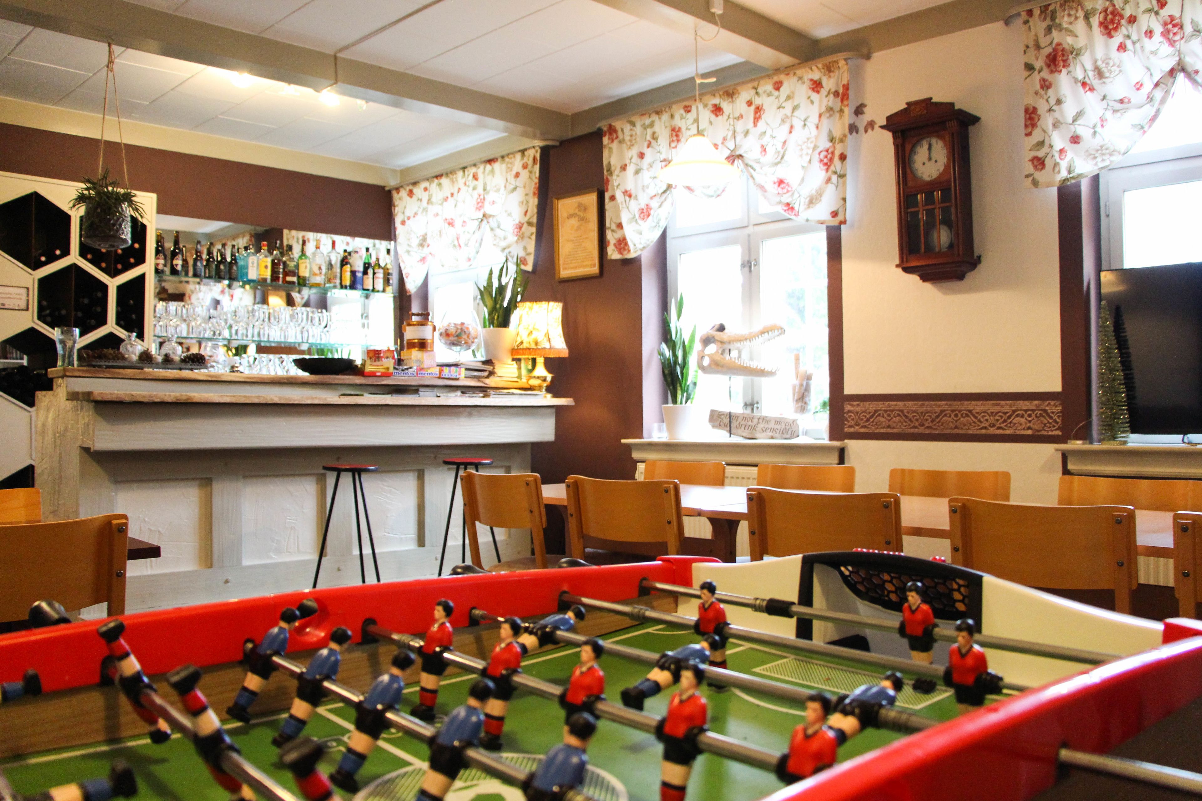 Zona de bar con mesa de fútbol de mesa, estanterías llenas de botellas y un reloj en la pared.