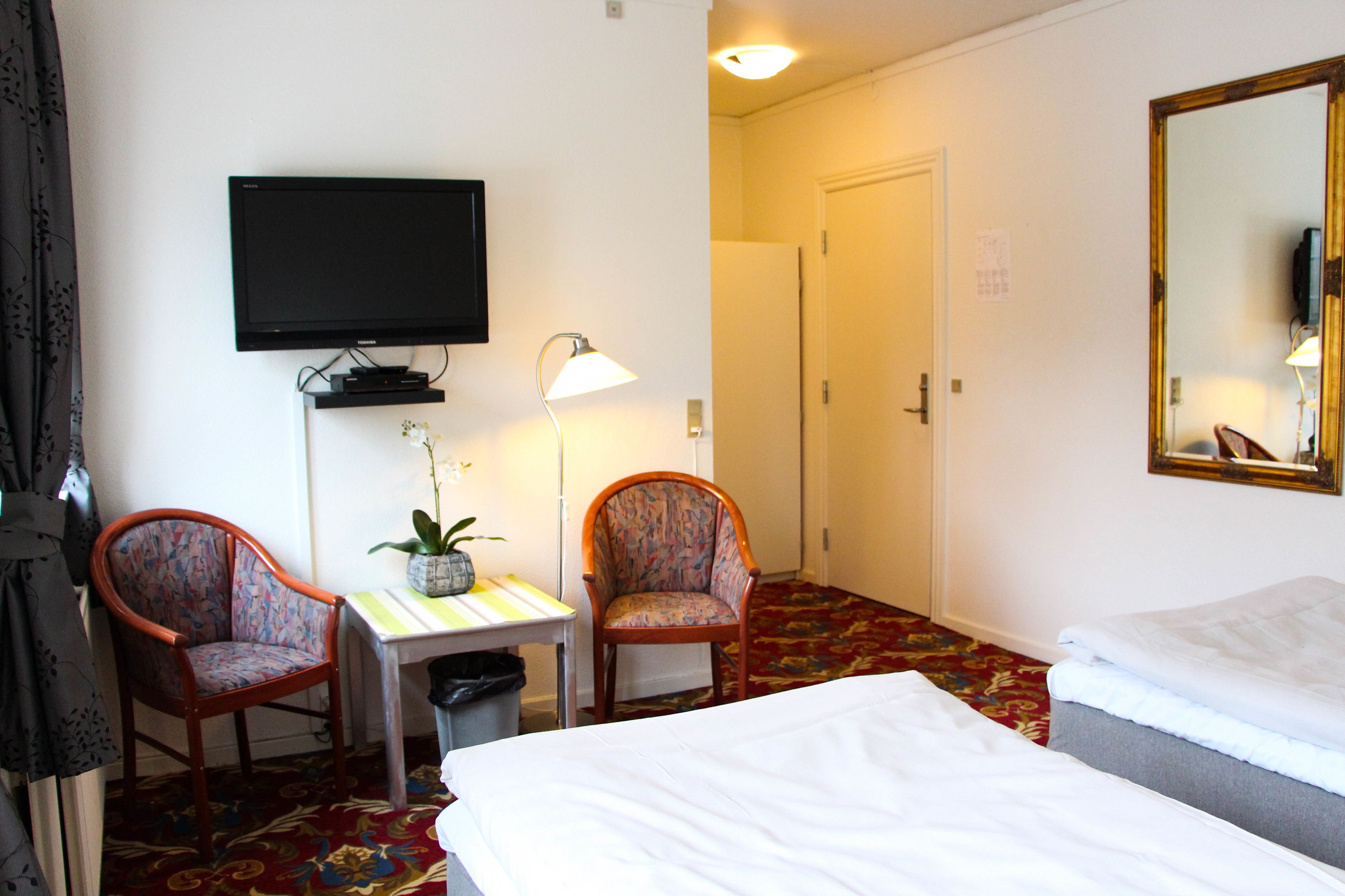 Et hotelværelse med to enkeltsenge, et fjernsyn ophængt på væggen, et skrivebord med lampe, to stole og et tæppe på gulvet.