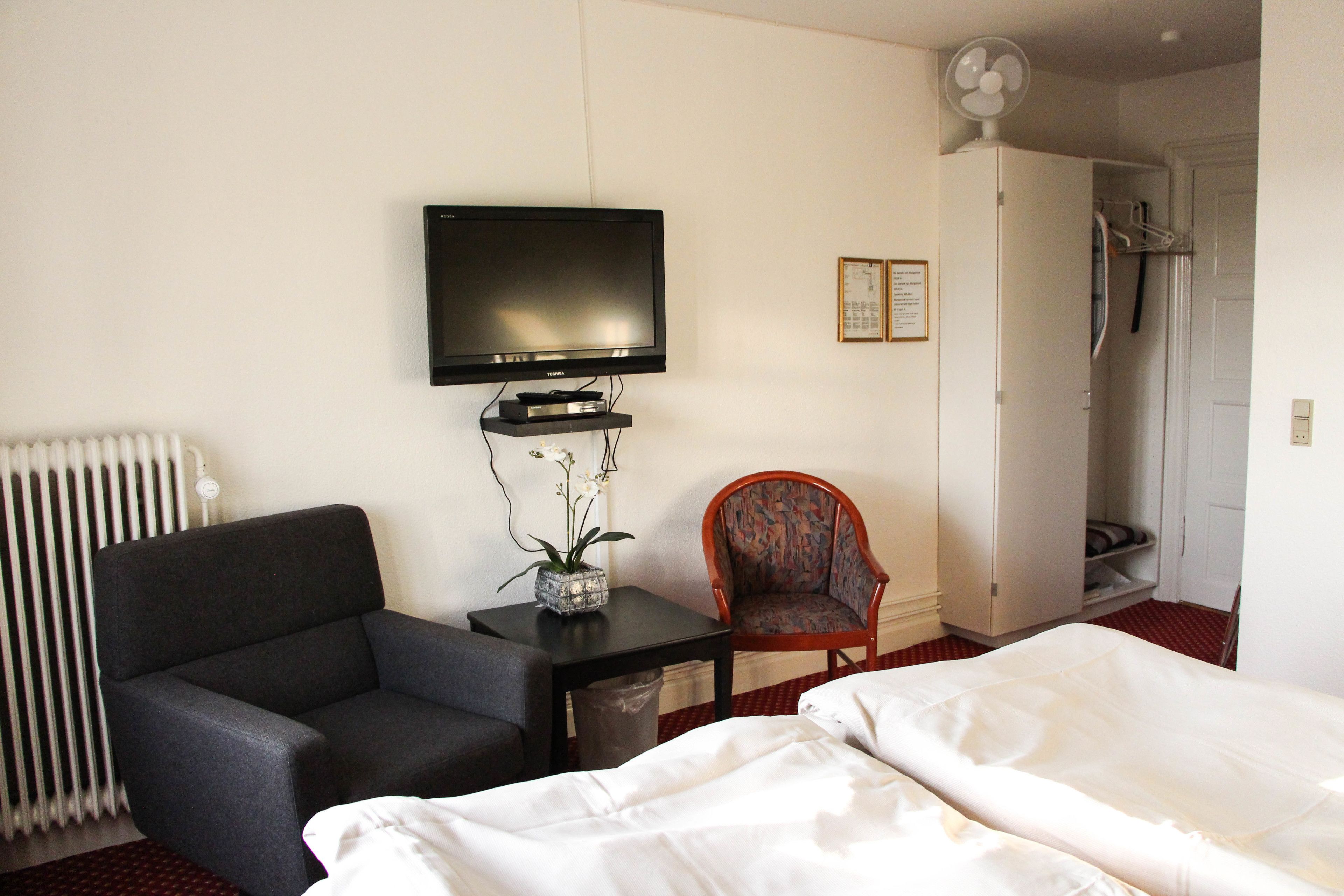 Habitación de hotel con cama, televisor sobre un mueble, una silla, una mesita con un florero y un radiador.