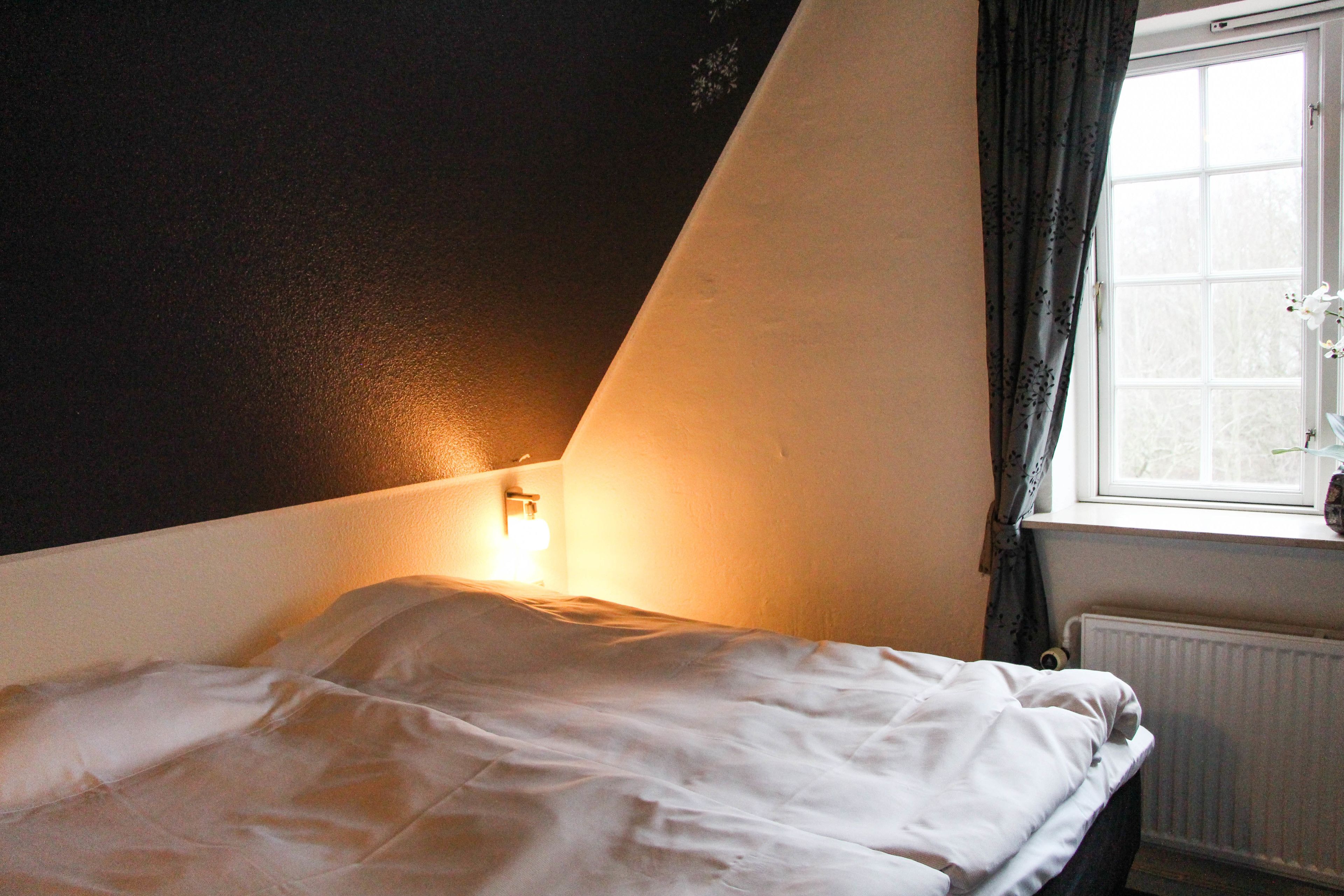 Un dormitorio con cama, ventana con cortinas y una lámpara fijada a la pared.