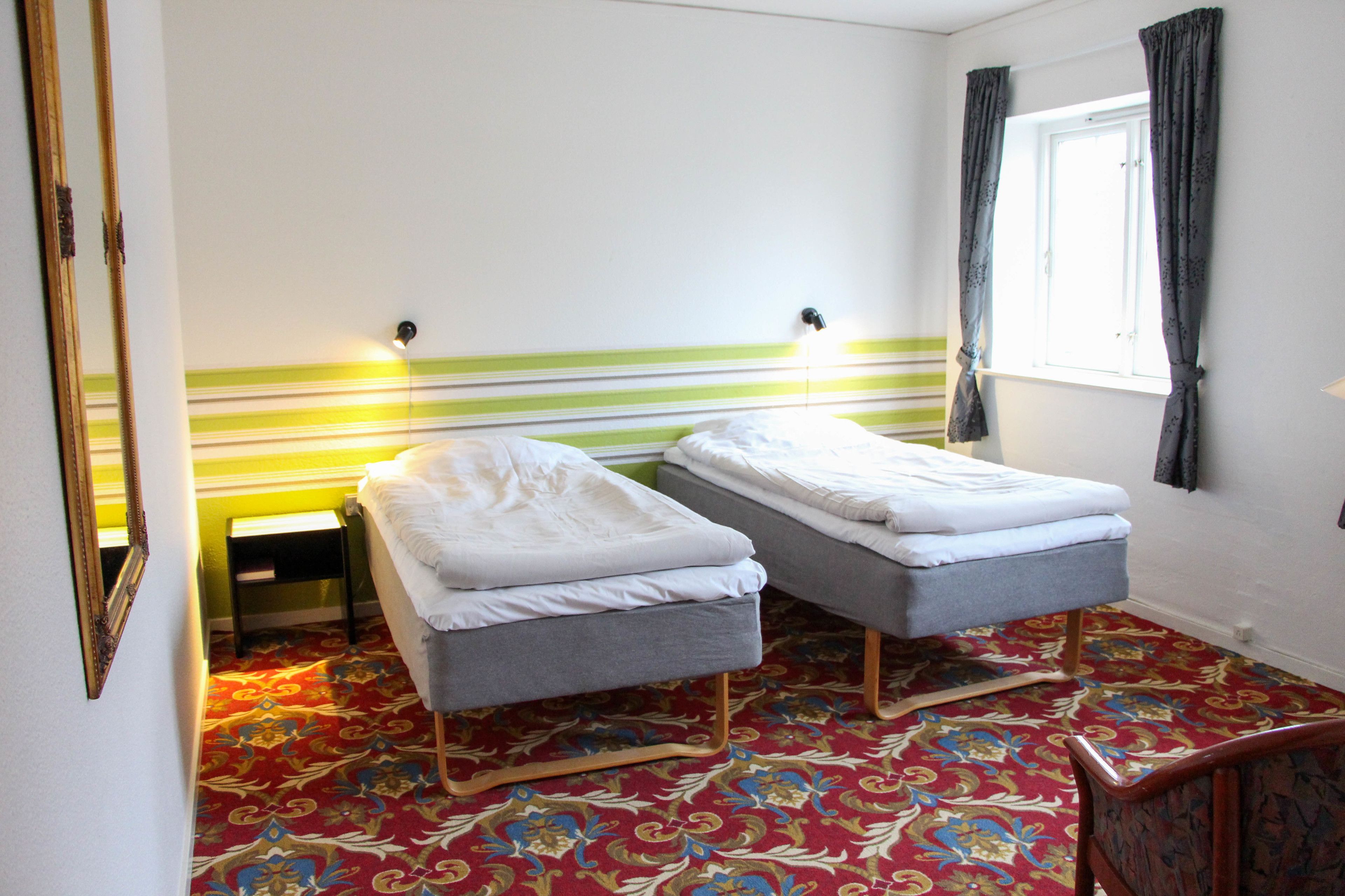 A room with two single beds, a small table, a mirror, and a window with curtains.