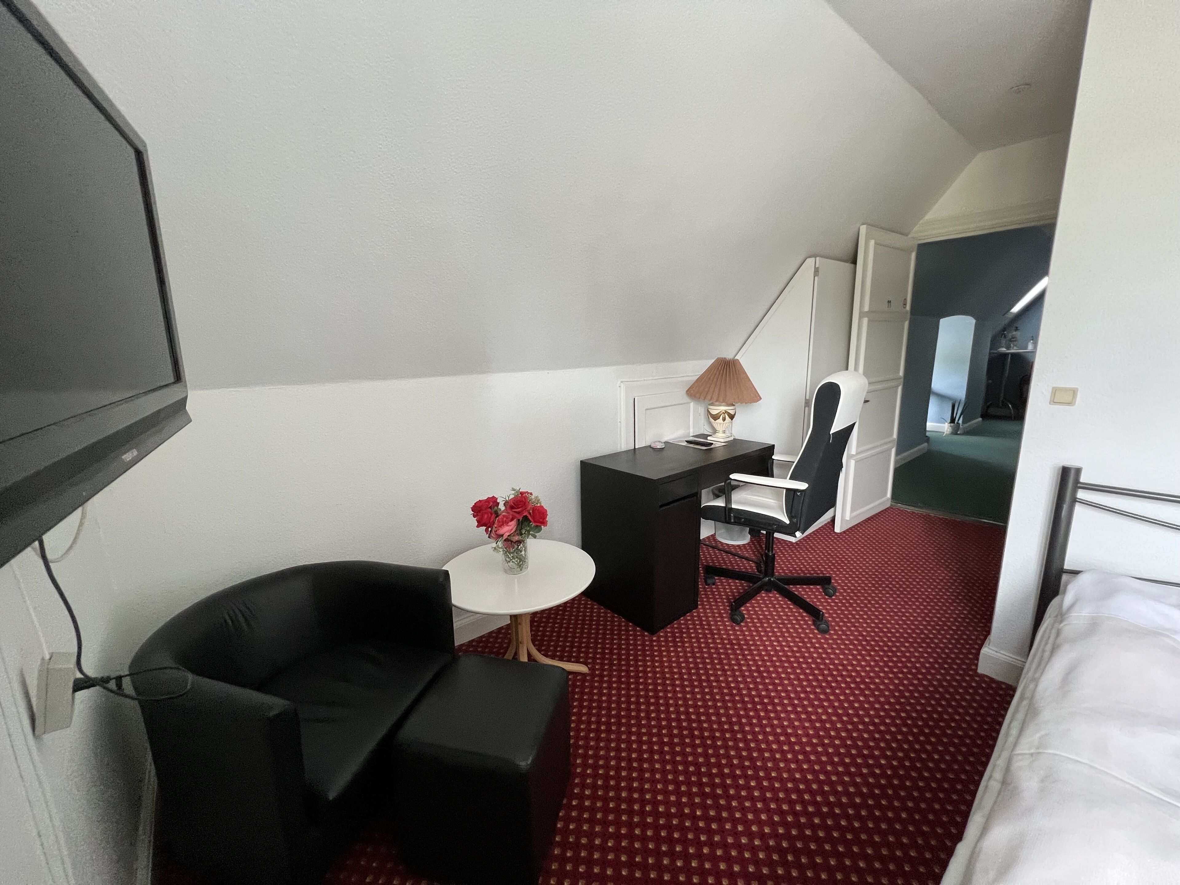 A hotel room with a bed, a desk, a chair, a small table with a vase of flowers, and a television.