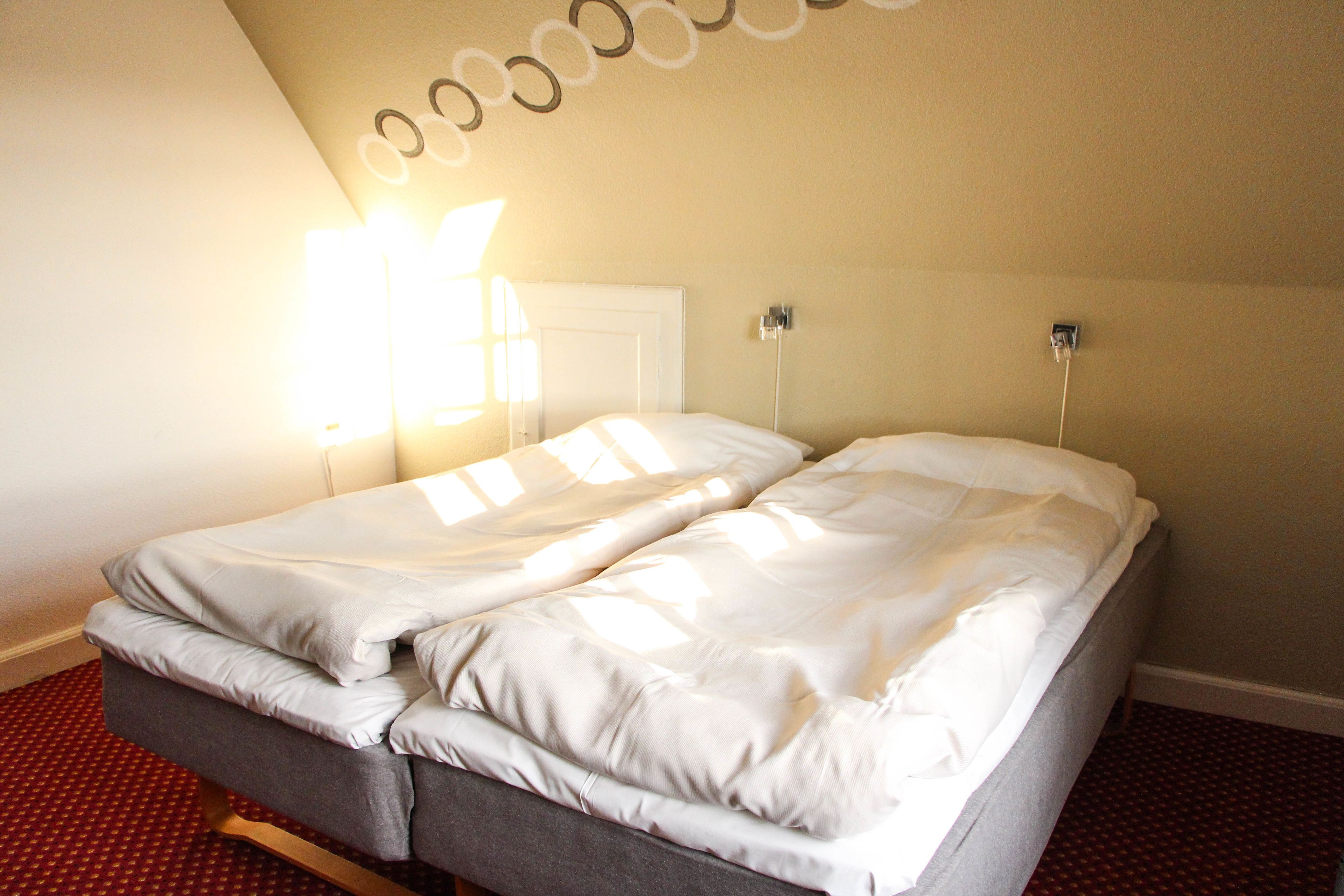 A double bed in a room with a red patterned carpet, a white door, and a wall-mounted light fixture.