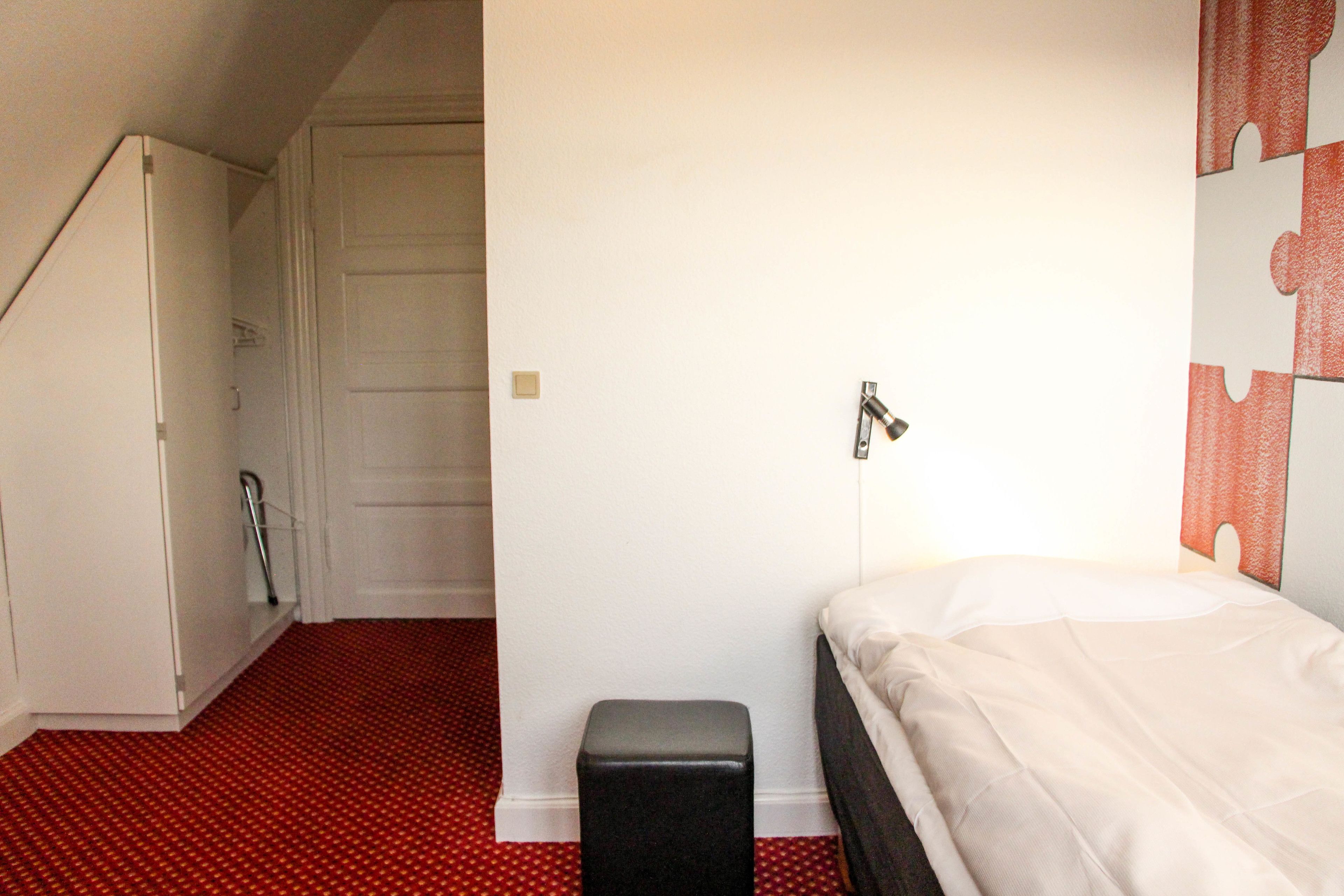 Un dormitorio con cama, un taburete pequeño, una puerta y una lámpara fijada a la pared.
