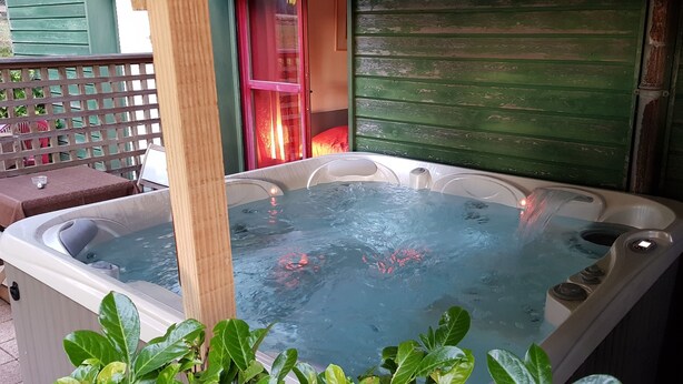 A hot tub with multiple seating areas, surrounded by greenery and a wooden fence. - Hôtel Le Pavillon (Cours)
