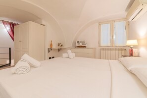 A hotel room with a large bed, a wardrobe, a radiator, a window with curtains, and a bedside table with a lamp.