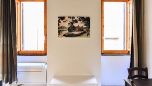 A room with two windows, a table, a chair, and a wall-mounted picture. - Spanish Steps Cozy Studio (Rome)