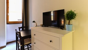A room with a TV on a white cabinet, a dining table with chairs, a potted plant, and a window with curtains. - Spanish Steps Cozy Studio (Rome)