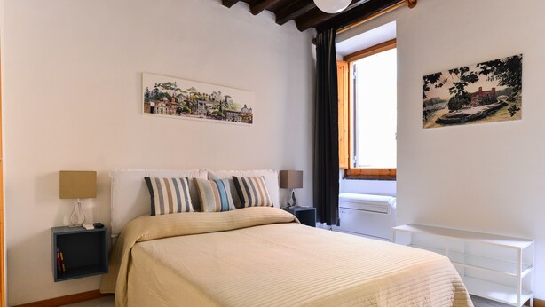 A neatly made bed with a beige quilt, two bedside tables with lamps, a white radiator, and two wall-mounted artworks depicting landscapes. - Spanish Steps Cozy Studio (Rome)