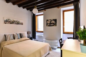 Un dormitorio con cama, escritorio y silla. Hay dos ventanas con cortinas, un ventilador de techo y un estante fijado a la pared.