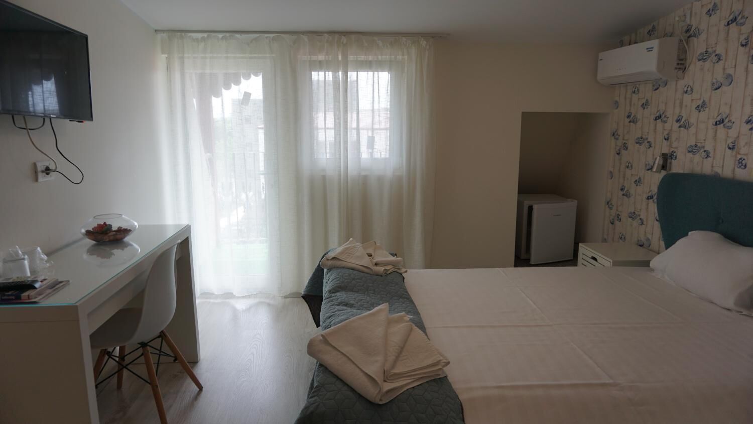 A hotel room with a bed, a desk with a chair, a television mounted on the wall, and a window with sheer curtains.