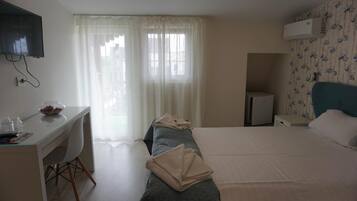 A hotel room with a bed, a desk with a chair, a television mounted on the wall, and a window with sheer curtains.