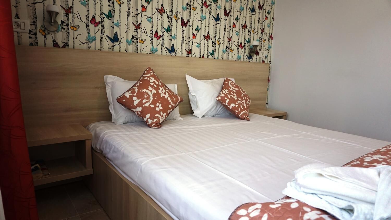 A neatly made bed with white bedding and two decorative pillows, a wooden headboard, and a wall with a floral pattern.