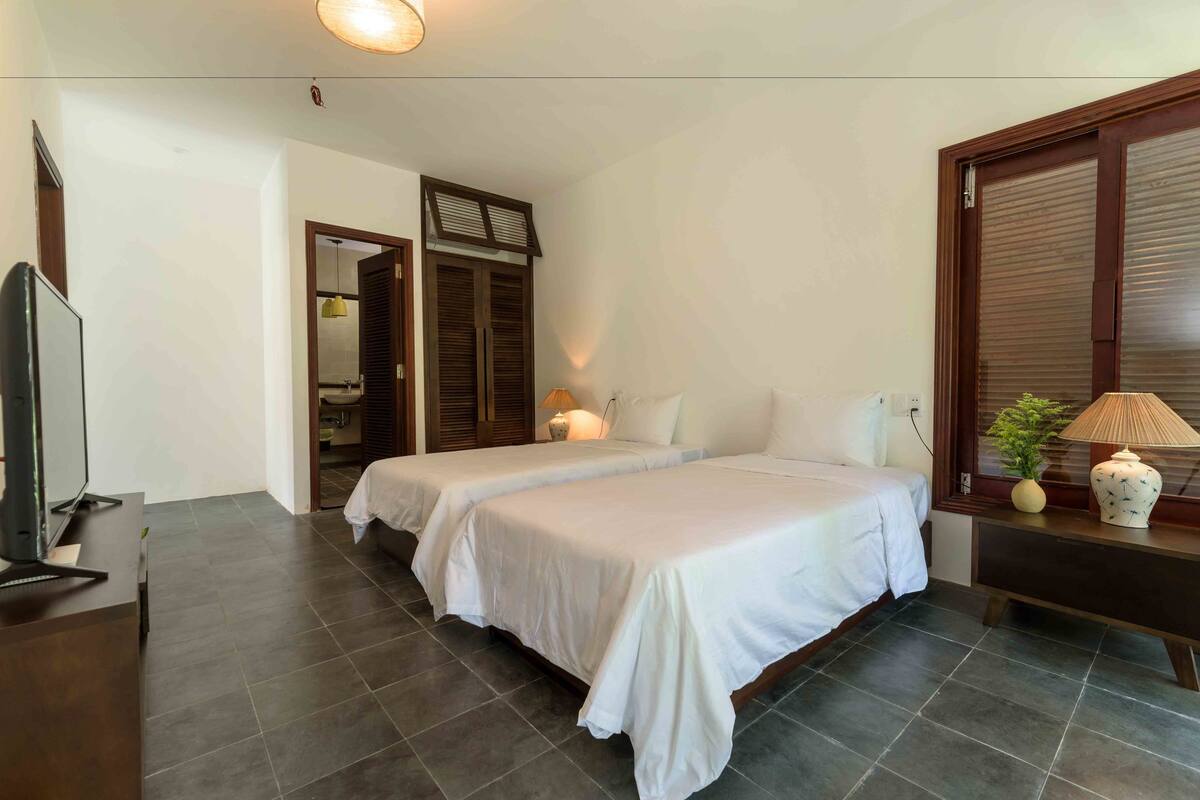 A hotel room with two beds, a television, a desk, and a window with shutters.