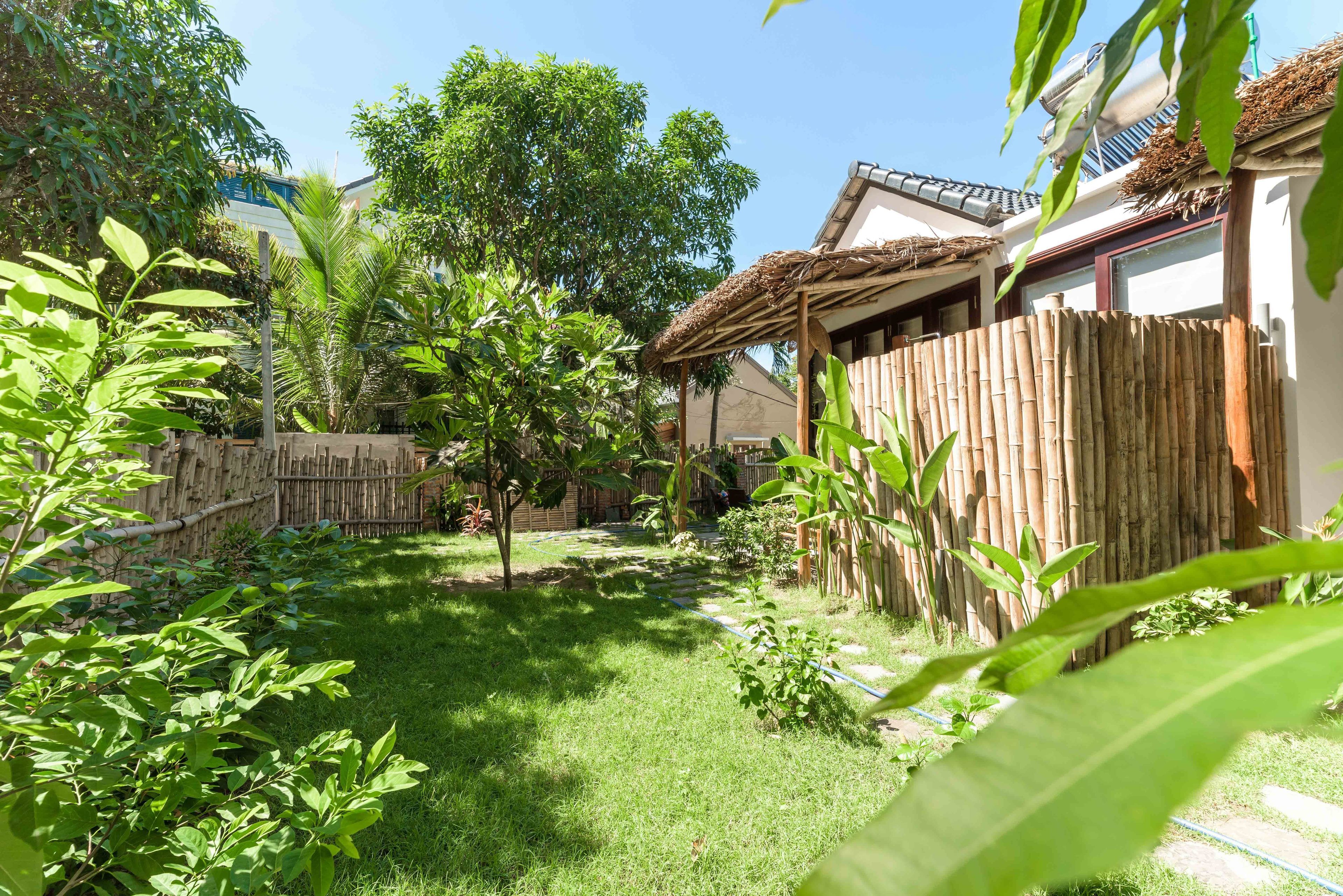 Un jardín con cerca de bambú, una estructura de techo de paja y abundante vegetación.