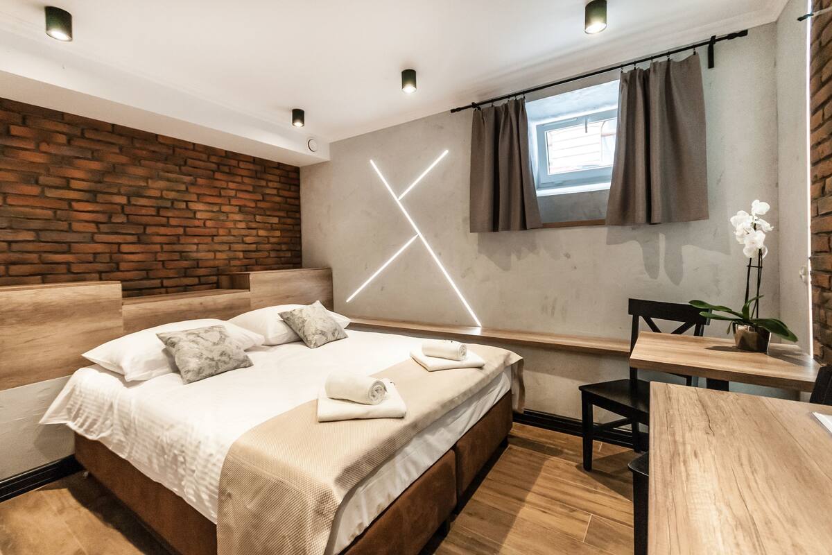 Quarto moderno com parede de tijolos, cama com roupa de cama branca, escrivaninha de madeira e planta em vaso.