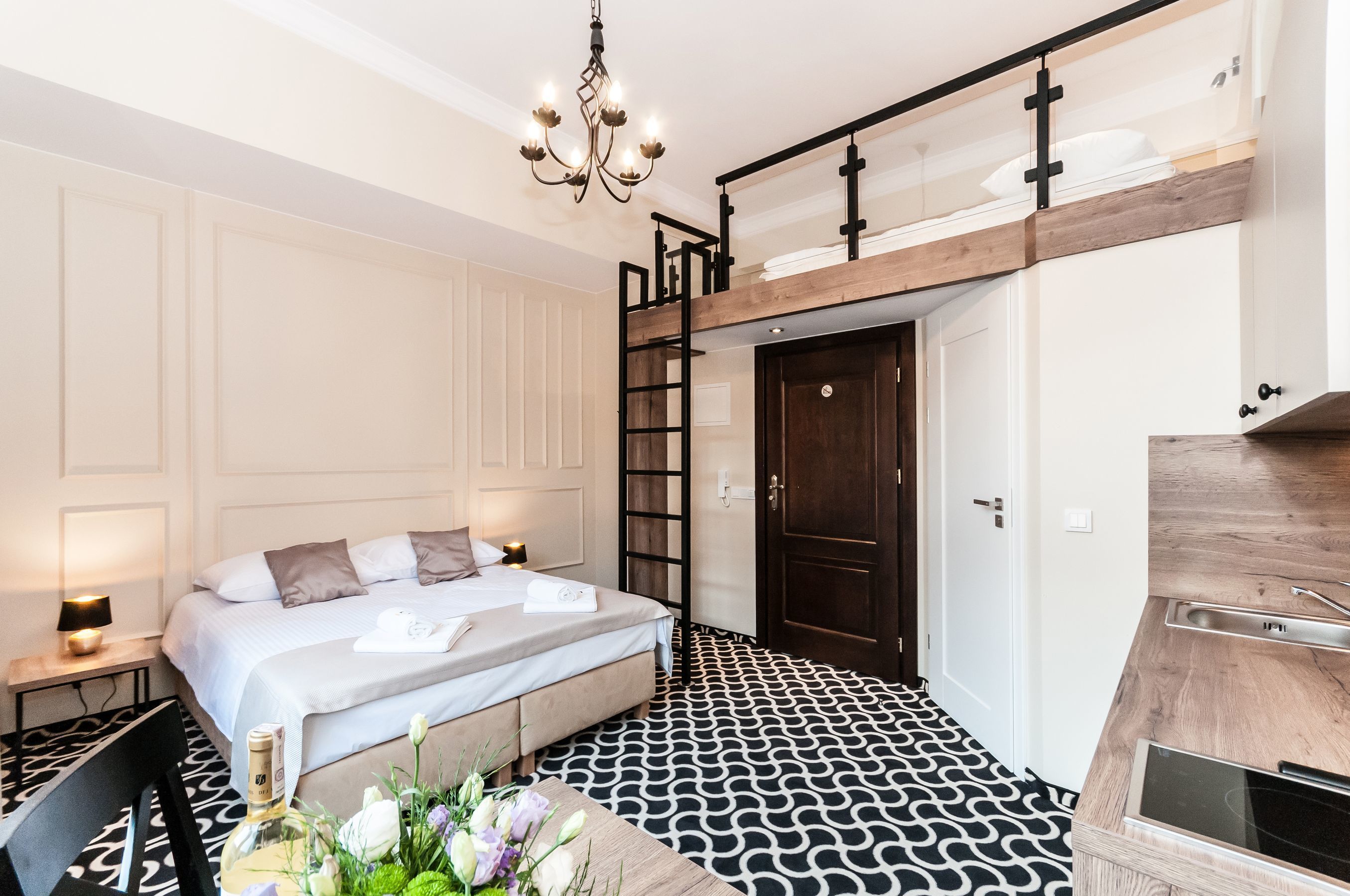 A modern bedroom with a bunk bed, a wooden ladder, a chandelier, a patterned floor, and a small kitchen area.