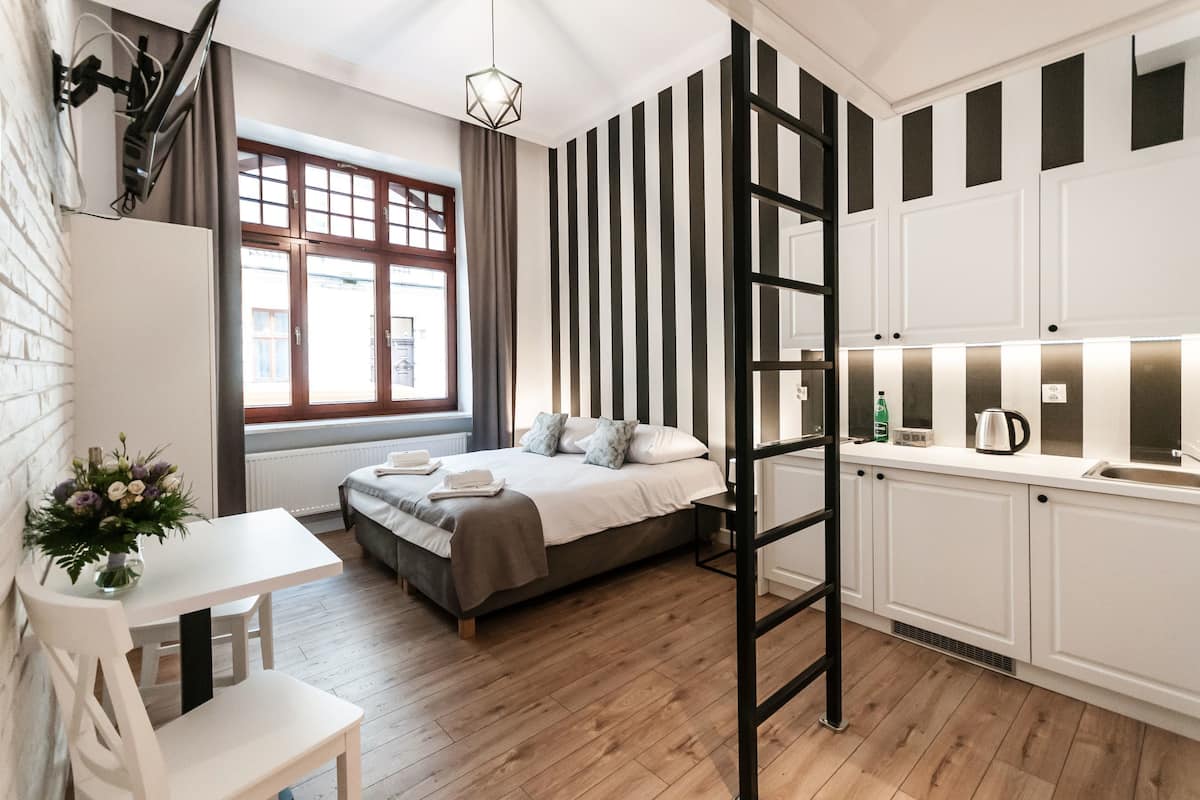 Quarto compacto com cama, mesa de cabeceira e um pequeno espaço de trabalho. Uma janela com cortinas oferece vista para o exterior.