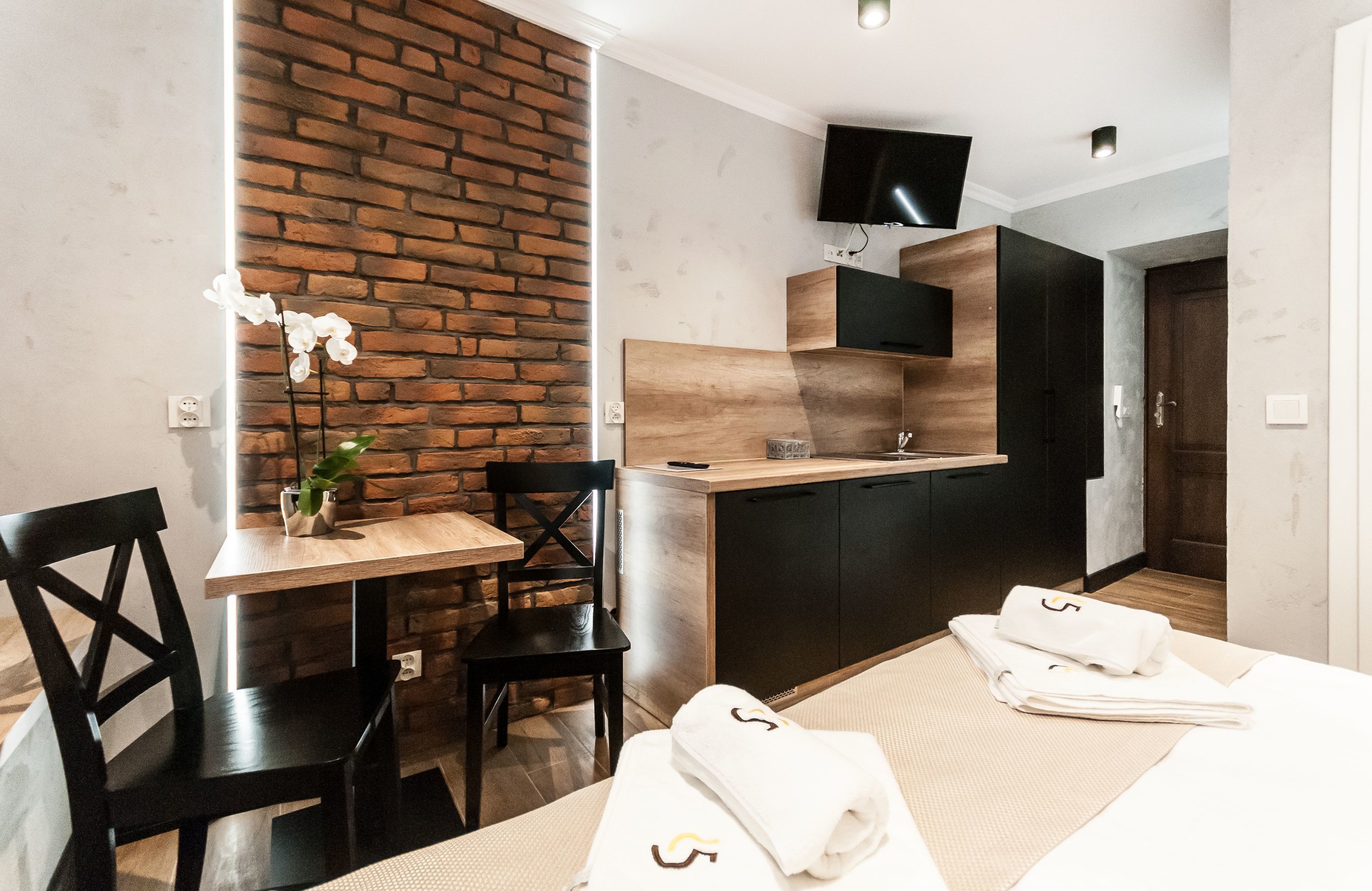 A compact hotel room with a brick accent wall, a small dining area, a kitchenette, and a flat-screen TV mounted above the counter.