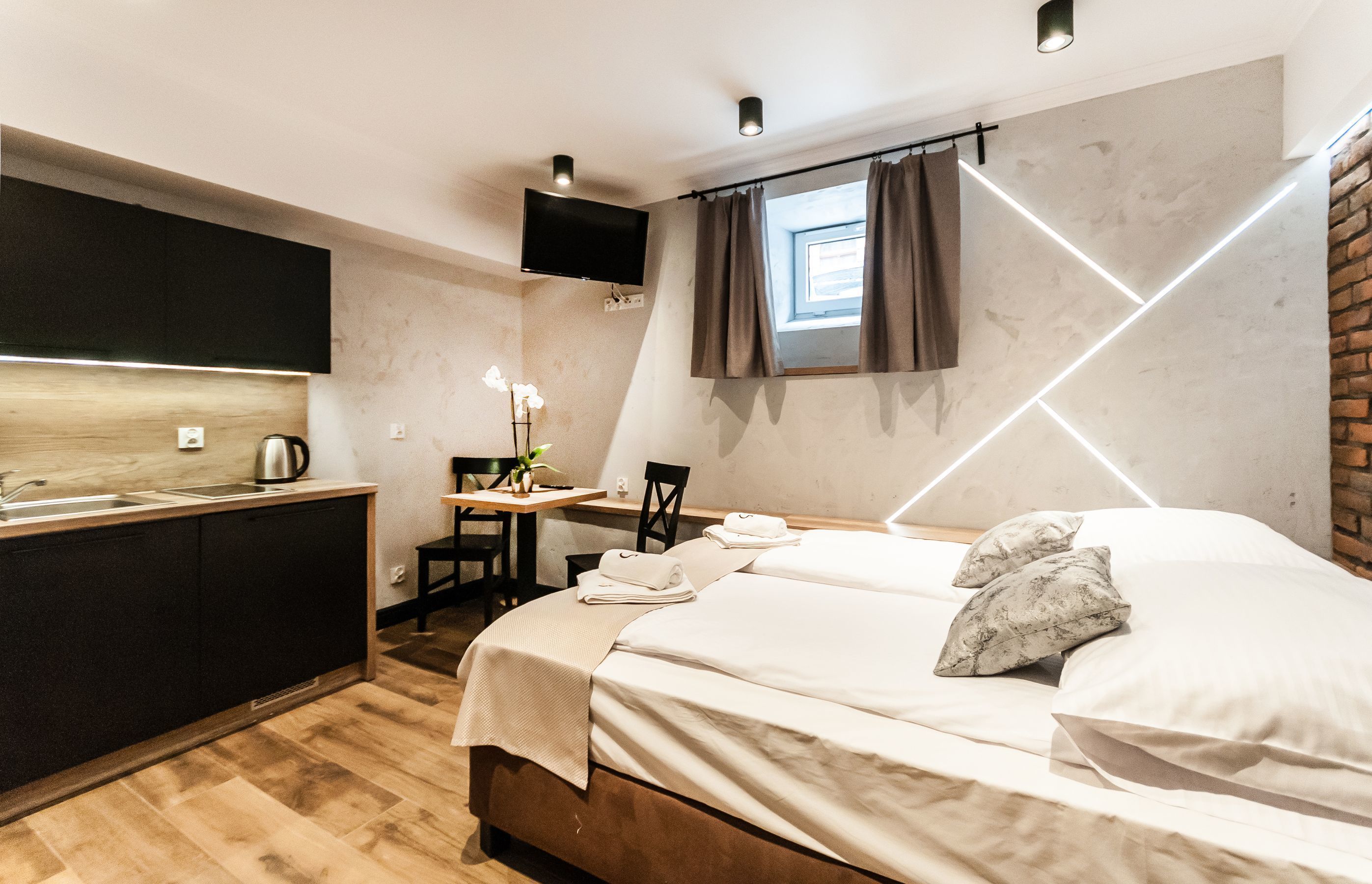 Ein modernes Hotelzimmer mit Holzboden, einem Bett mit weißen Leinen, einem Schreibtisch mit Stuhl, einem kleinen Tisch mit Pflanze, einem an der Wand montierten Fernseher und einem Fenster mit Vorhängen.