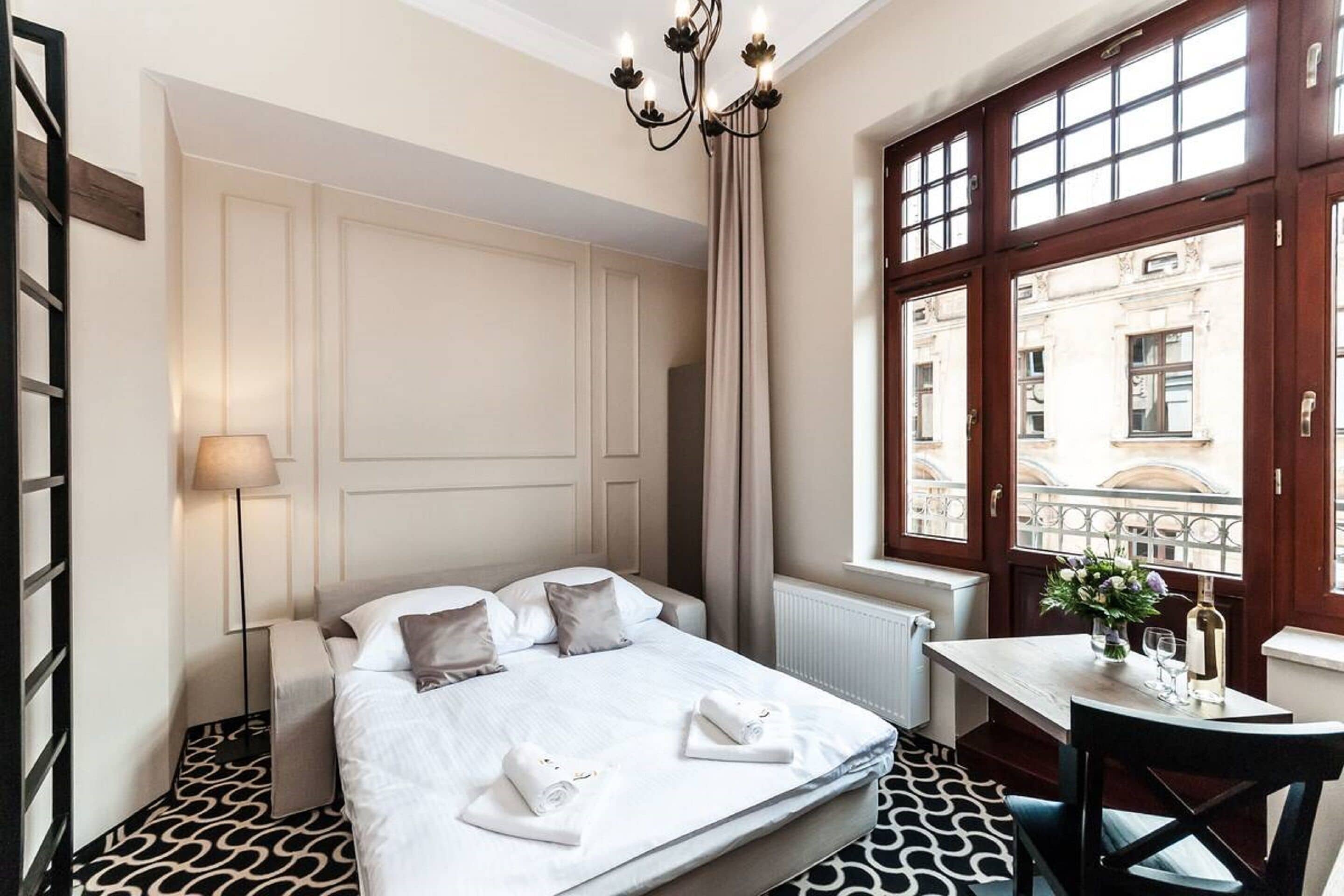 A hotel room with a bed, a table with a vase and wine bottle, a window with a view of buildings, and a chandelier.