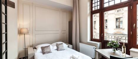 A hotel room with a bed, a table with a vase and wine bottle, a window with a view of buildings, and a chandelier.