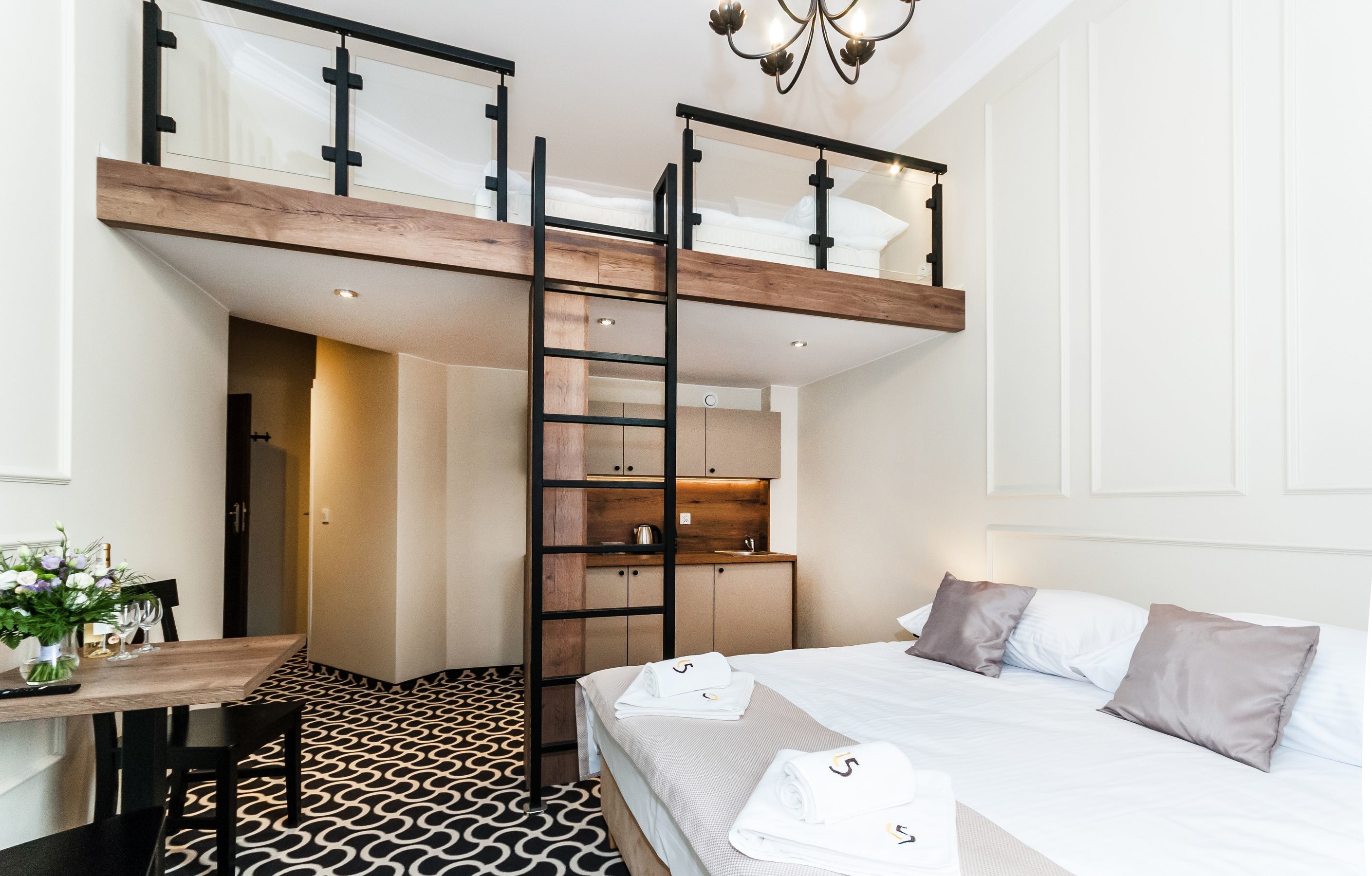 A hotel room with a loft bed, a desk with a chair, a patterned floor, and a kitchenette area.