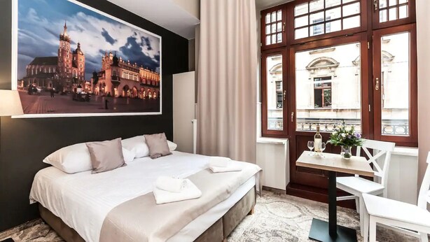 Una habitación de hotel con una cama grande, un escritorio con una silla, una ventana con vistas a un edificio y un cuadro de un paisaje urbano.