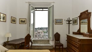 A bedroom with a bed, a wooden dresser, a chair, a desk with a lamp, and a window with a view of buildings. - Rome at Your Feet Apartment (Rome)