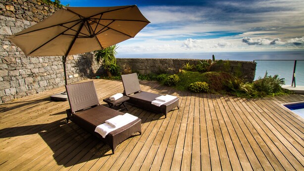 A wooden deck with two lounge chairs and a large umbrella, overlooking a pool and a stone wall.