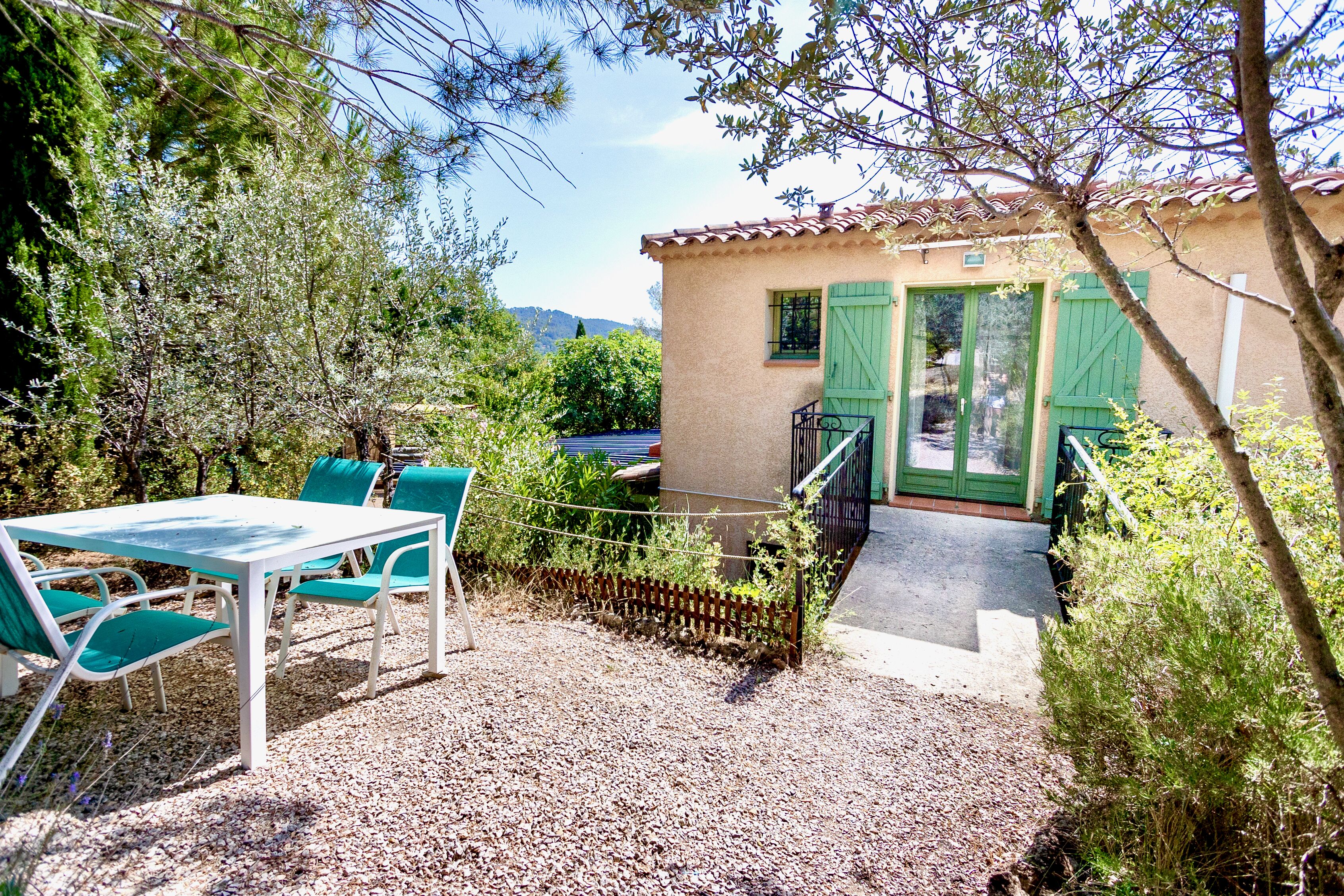 A patio with a table and chairs, a small building with green shutters, and a gravel ground.