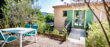 A patio with a table and chairs, a small building with green shutters, and a gravel ground.