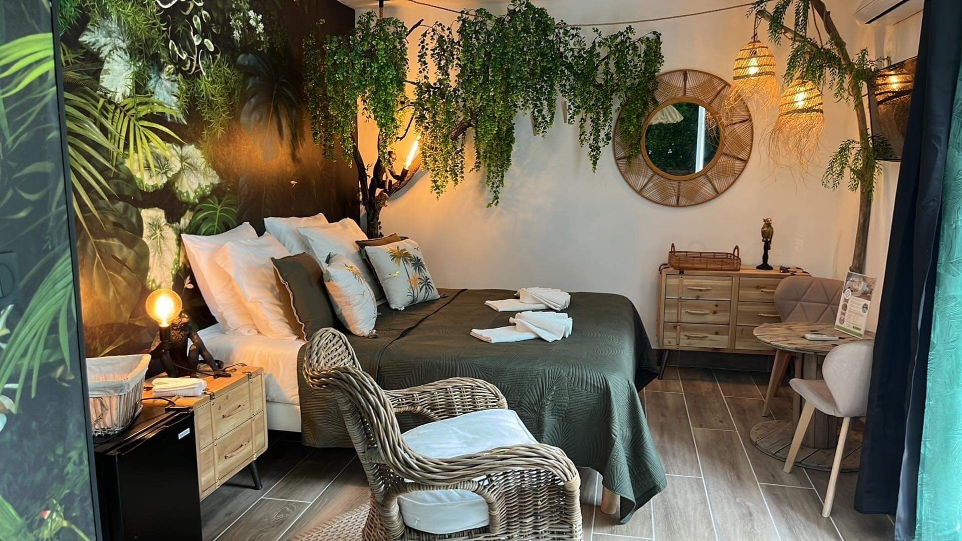 A bedroom with a bed, nightstand, wicker chair, dresser, and hanging plants.