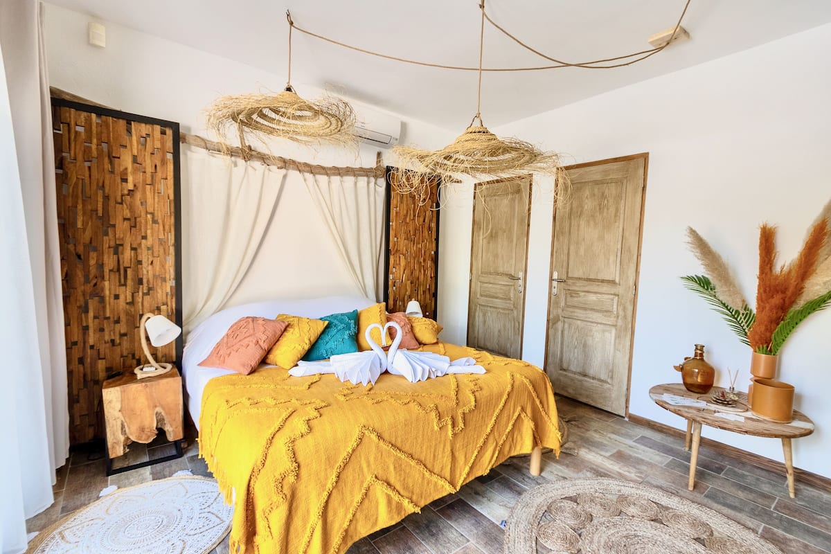 A well-lit bedroom with a bed, a bedside table, a hanging light fixture, and a small round table with a vase.