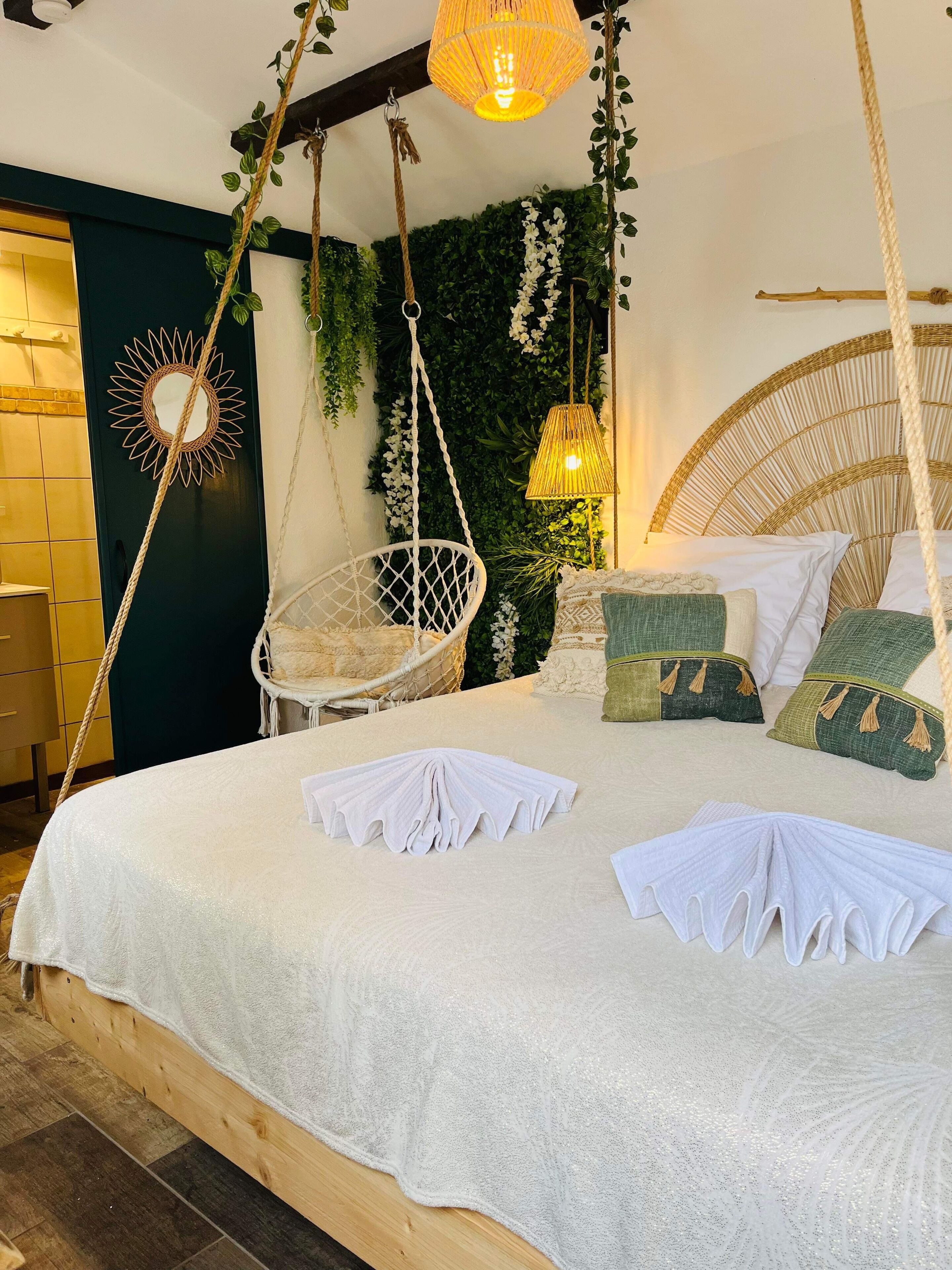 A bed with a white bedspread, a hanging chair, and a wall with green foliage.